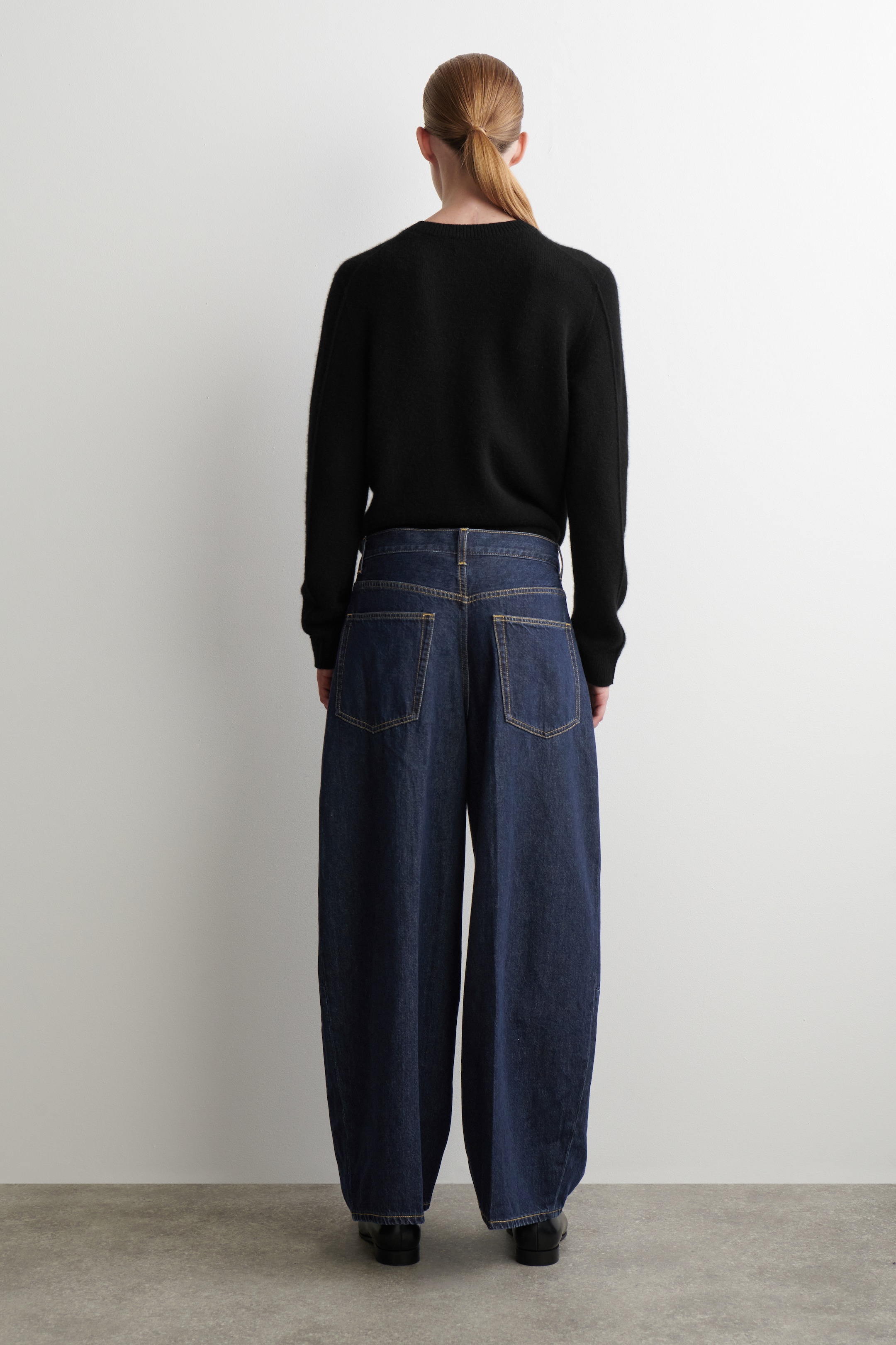 Visualizza immagine più grande: Un maglione nero a girocollo e jeans blu scuro a gamba larga vengono mostrati da dietro. Il maglione presenta una vestibilità rilassata, mentre i jeans mostrano una silhouette a vita alta e gamba larga con cuciture gialle a contrasto, passanti per cintura e due grandi tasche posteriori applicate.
