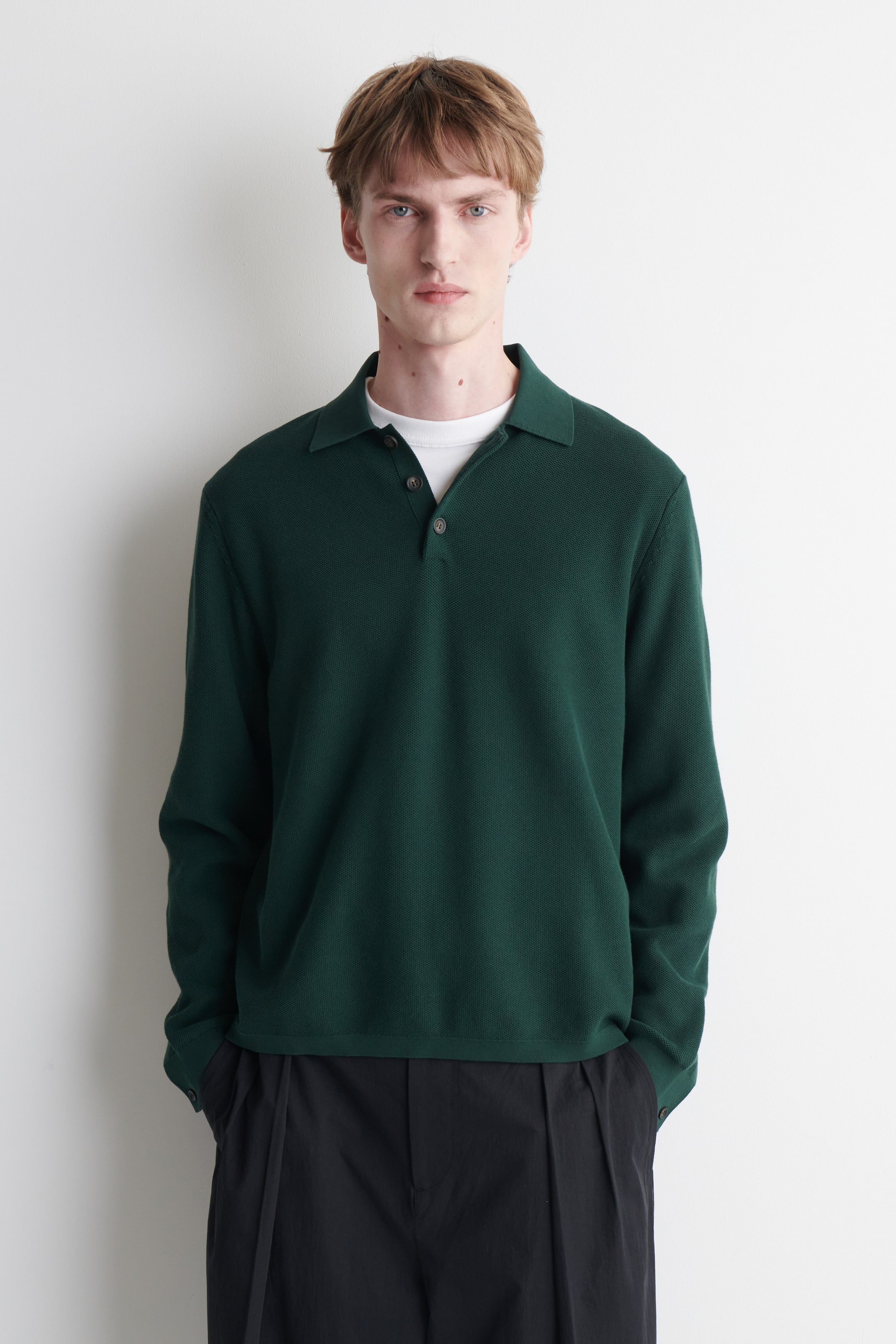 Agrandir l'image: Un polo à manches longues vert foncé est porté, avec un col ouvert et une patte de boutonnage à deux boutons avec des boutons foncés. La chemise présente un tricot texturé et est superposée sur un t-shirt blanc à col rond.