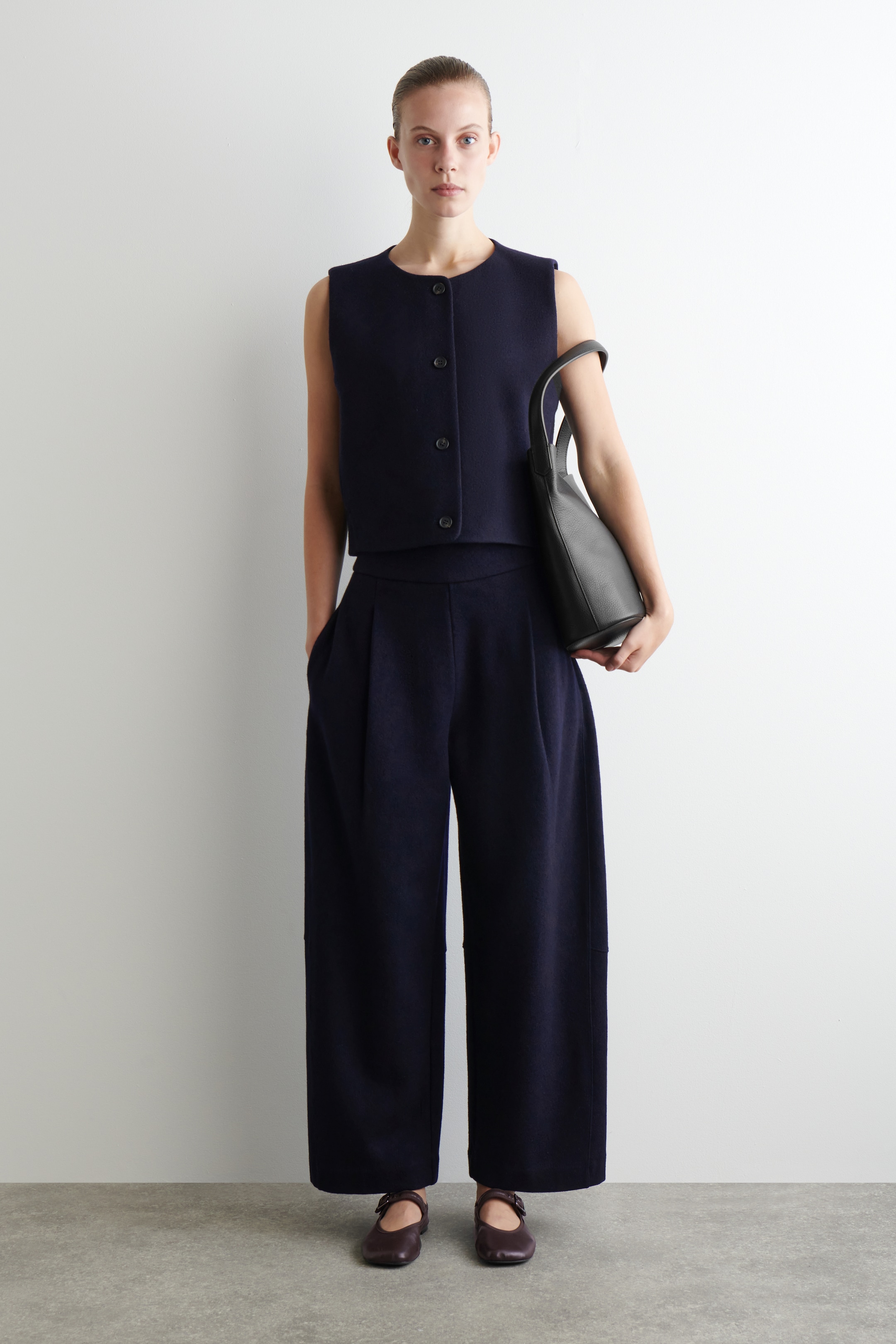 Ingrandisci l'immagine: Un completo a due pezzi di un blu navy scuro viene presentato da un'angolazione frontale, caratterizzato da un gilet senza maniche, corto, con scollo arrotondato e chiusura frontale con bottoni, abbinato a pantaloni a vita alta e gamba larga con pieghe frontali. L'outfit è completato da una borsa tote in pelle nera, tenuta di lato, e da ballerine Mary Jane di un rosso scuro.