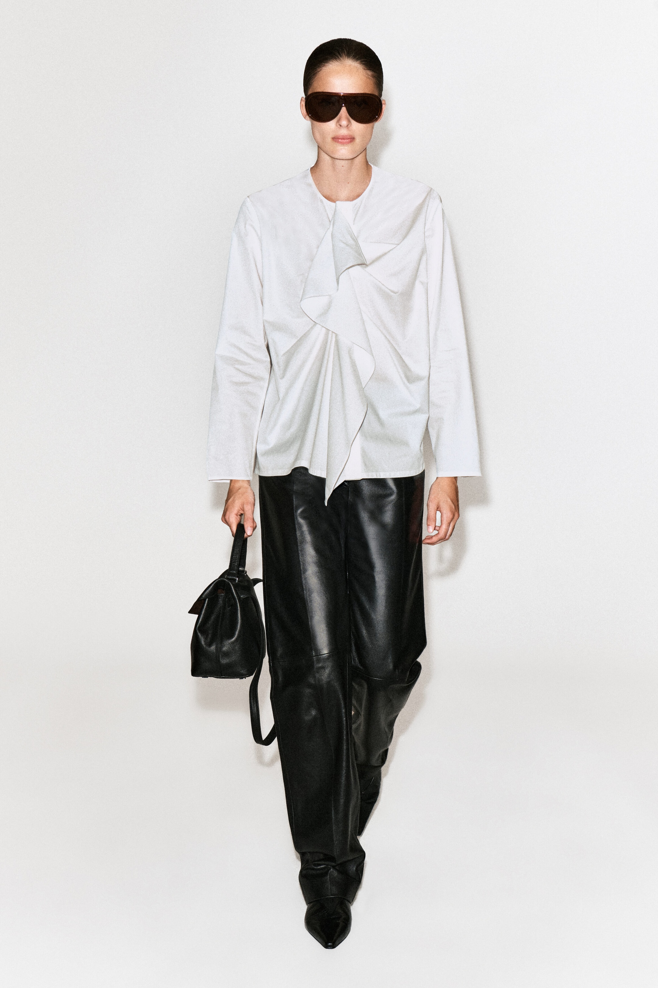 The model is wearing a white cotton blouse with long sleeves, a crew neckline, and an asymmetrical cascading ruffle at the front. The blouse is styled with black leather wide-leg trousers and a black leather handbag held in one hand.