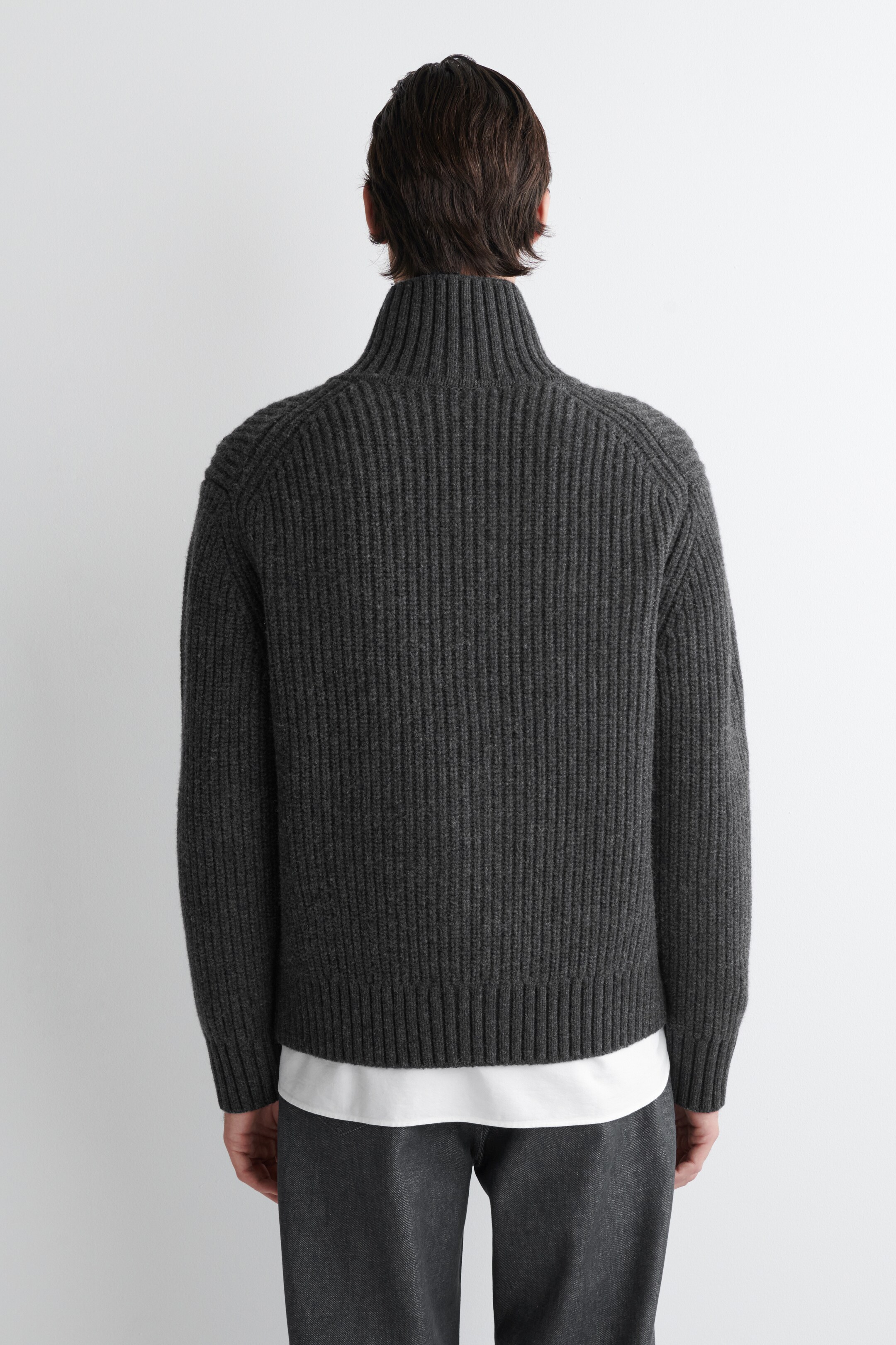 Agrandir l'image: Un pull en tricot côtelé gris foncé est présenté de dos. Il présente un col montant, des manches raglan et un ourlet côtelé. Un sous-vêtement blanc est visiblement superposé sous le pull.
