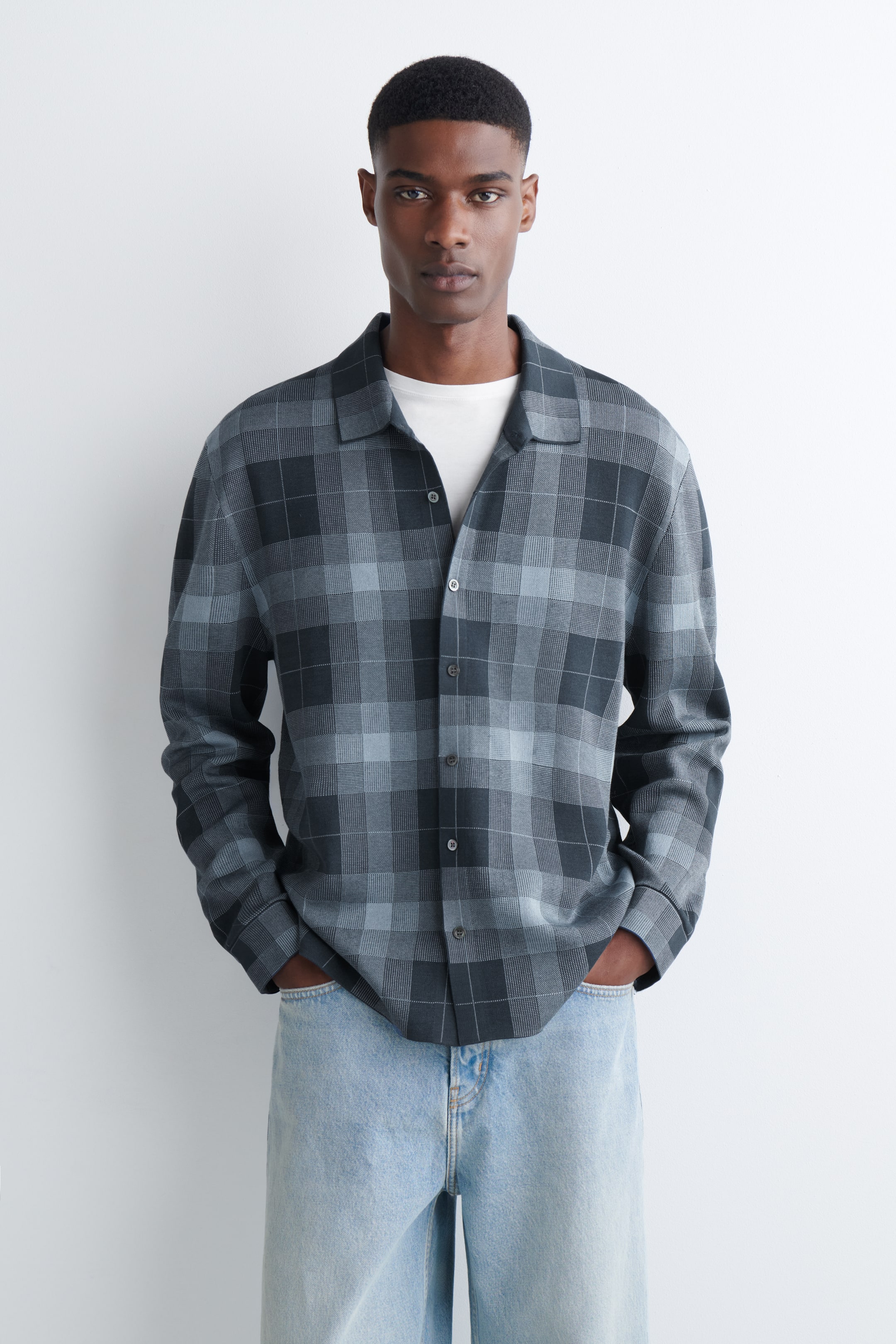 Grösseres Bild ansehen: Ein graues Karohemd wird von vorne gezeigt, offen getragen über einem weißen T-Shirt, mit einem Camp-Kragen und langen Ärmeln. Es zeigt ein Karomuster in verschiedenen Schattierungen von Anthrazit und Hellgrau und wird gestylt mit hell gewaschenen Jeans präsentiert.