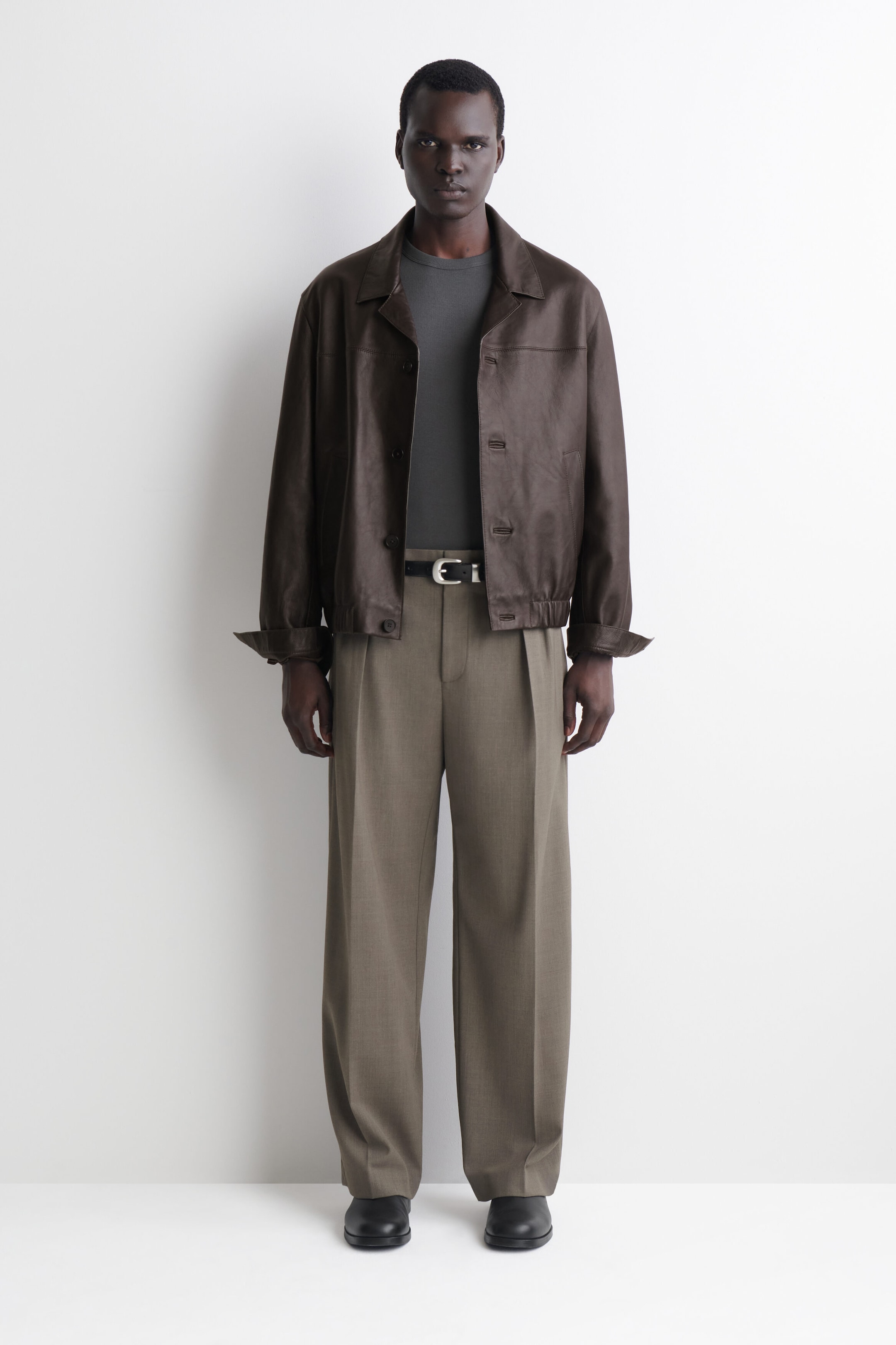 A slim ribbed cotton T-shirt in dark grey is shown from the front, featuring a crew neck and a fine ribbed texture. The T-shirt fits closely to the body, tucked into belted trousers and layered underneath an open brown leather jacket.