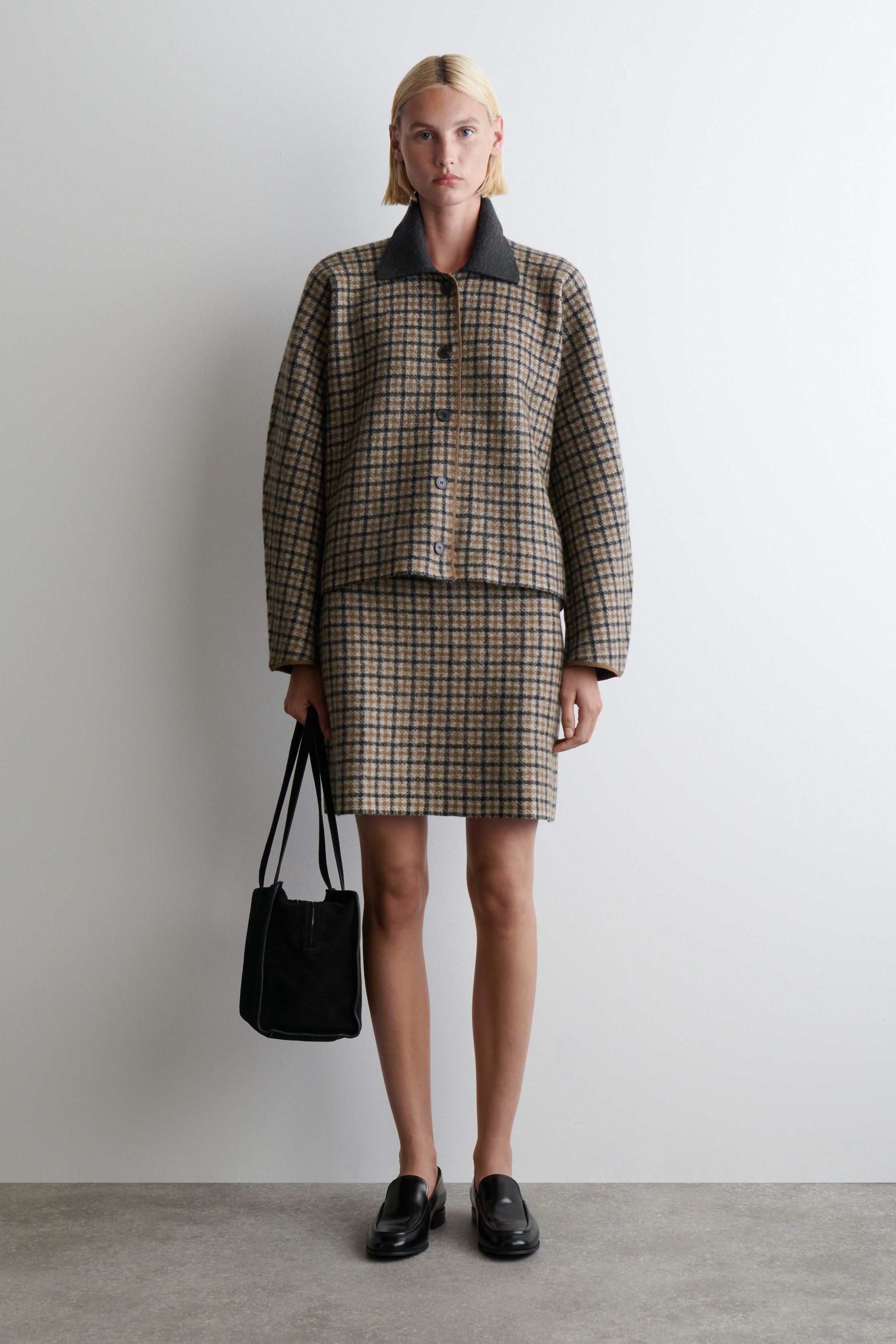 Grösseres Bild ansehen: Eine braune, schwarze und beige karierte Jacke und ein passender Minirock werden von vorne präsentiert. Die Jacke hat einen dunkelgrauen Umlegekragen und einen Knopfverschluss mit einem vertikalen braunen Besatz. Eine schwarze strukturierte Handtasche und schwarze Loafer vervollständigen das gestylte Ensemble.