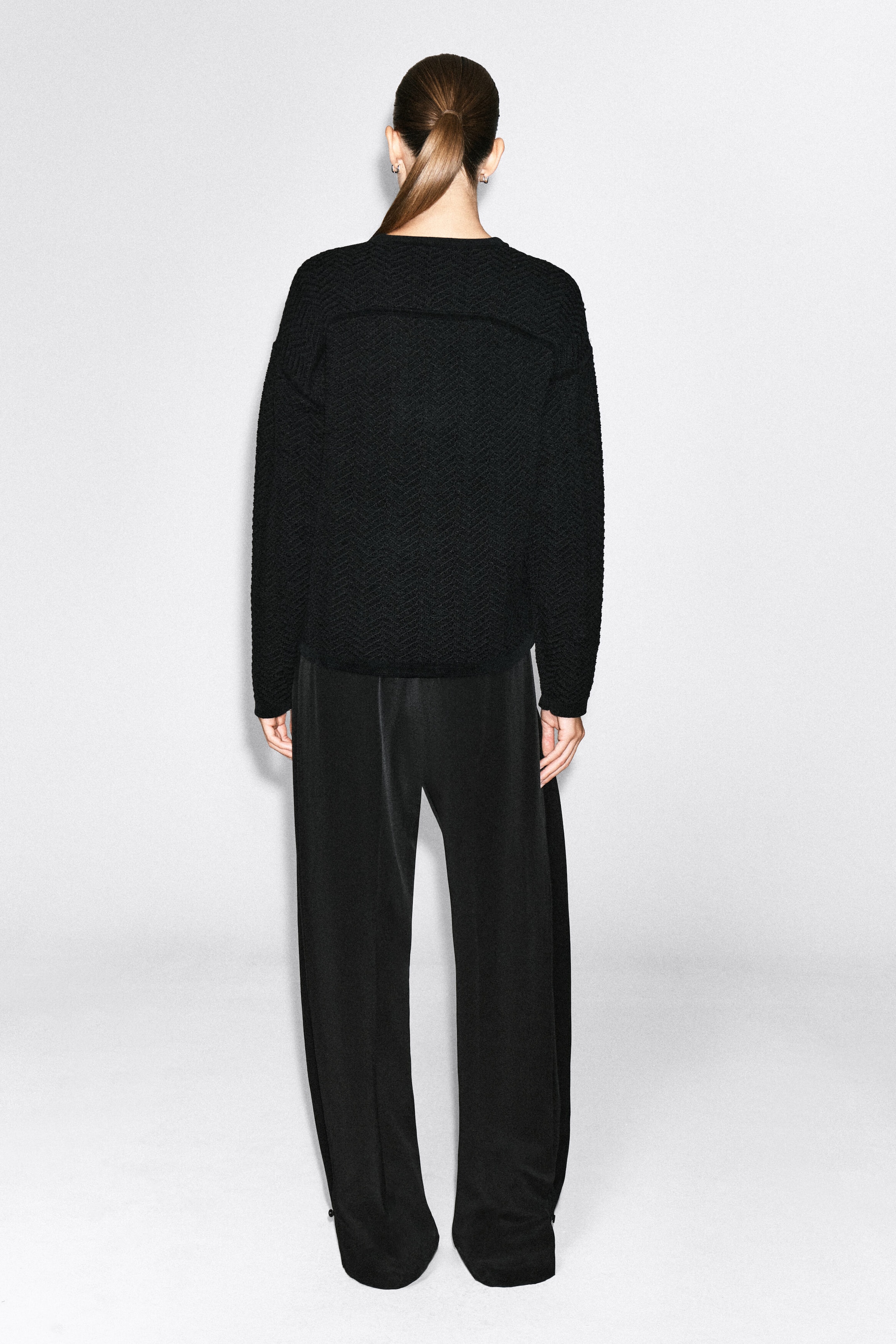 A black merino wool chenille crew-neck sweater with a textured chevron pattern is worn by the model, shown from the back. The long-sleeved sweater is paired with matching black wide-leg trousers. A black merino wool chenille crew-neck sweater with a textured chevron pattern is worn by the model, shown from the back. The long-sleeved sweater is paired with matching black wide-leg trousers.