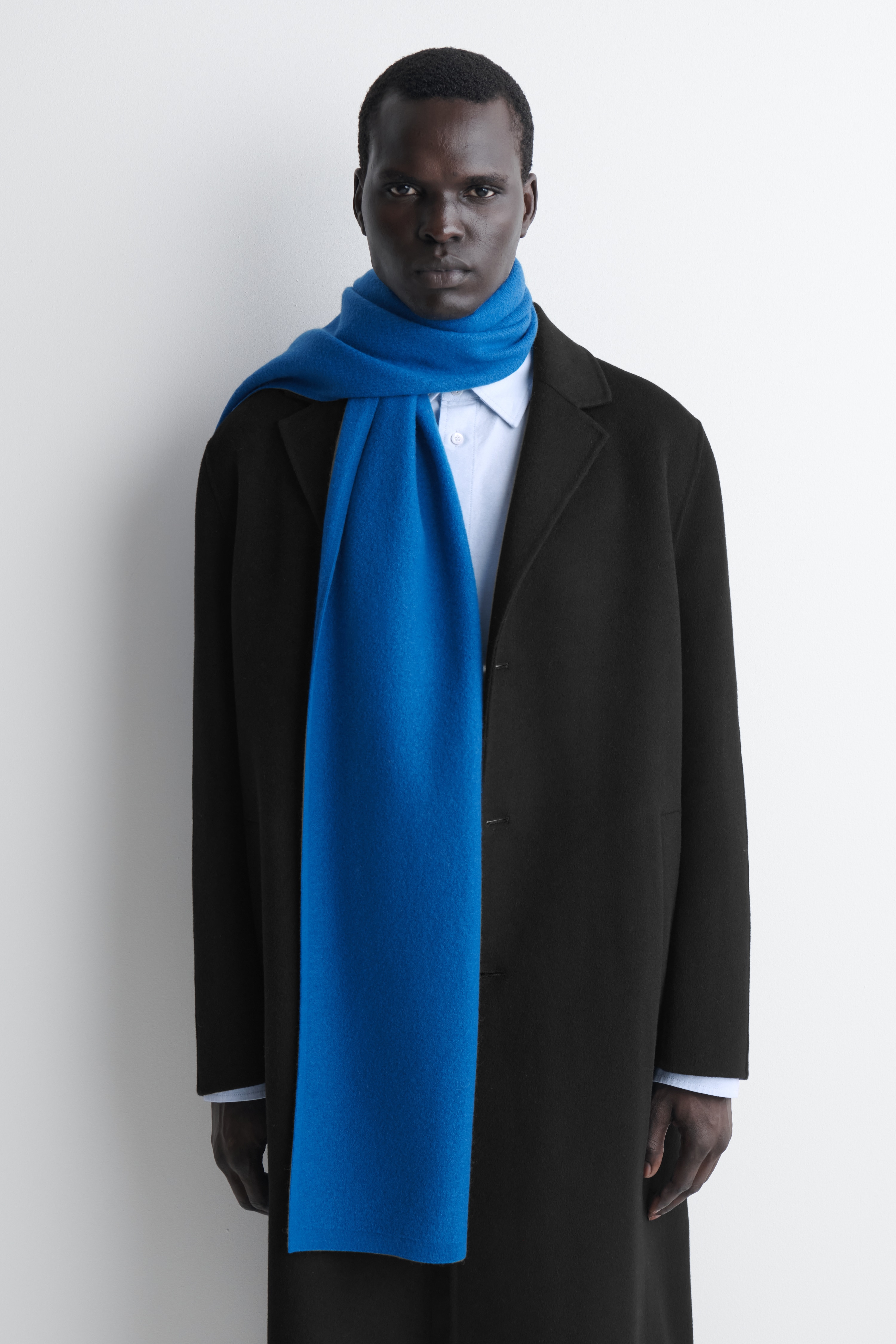 En blÃ¥ scarf i kokt ull ligger plant i en vikt rektangulÃ¤r form och visar upp sin strukturerade yta samt en liten vit varumÃ¤rkesetikett i det nedre vÃ¤nstra hÃ¶rnet.