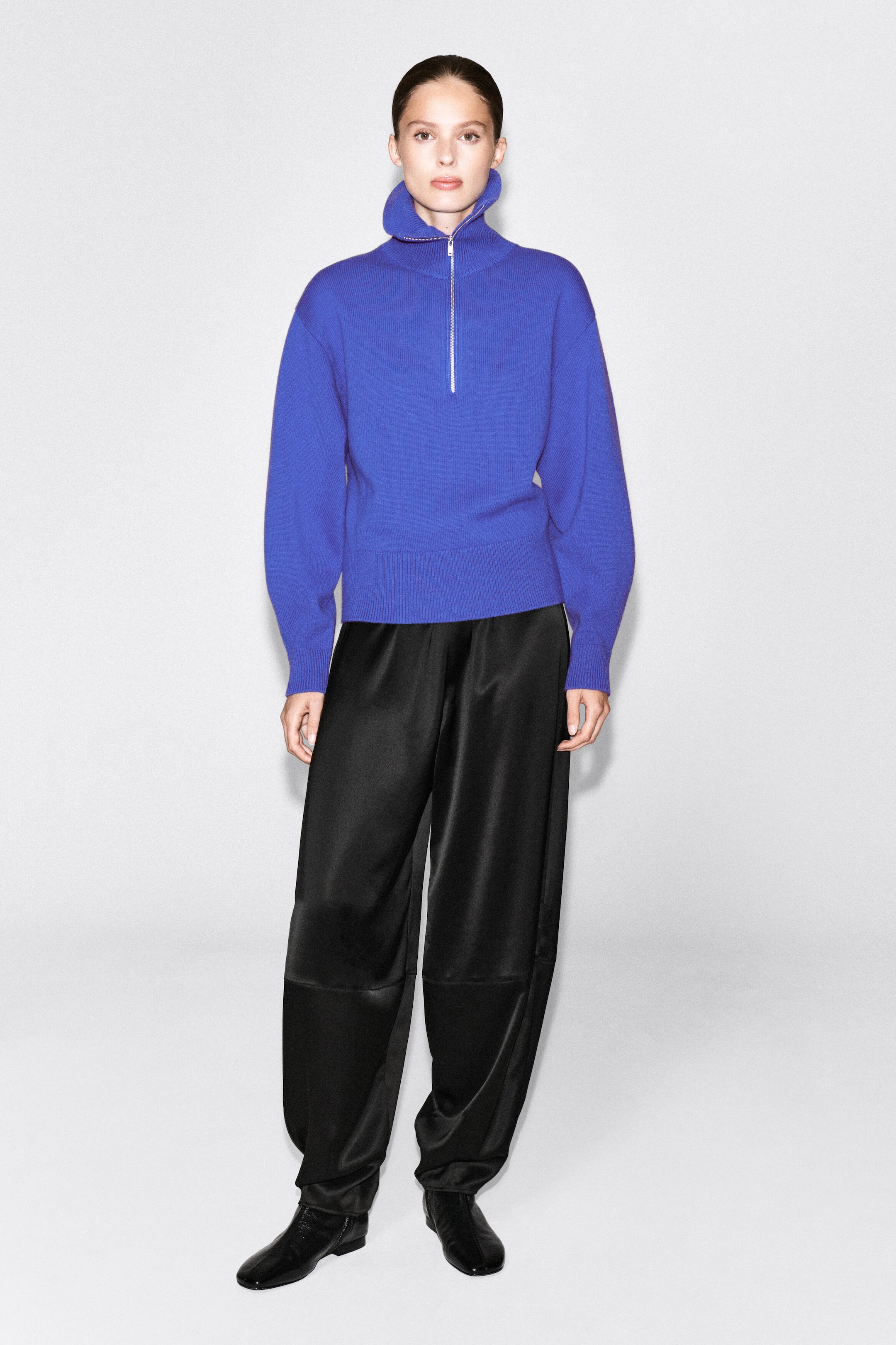 The model is wearing a bright blue knitted sweater with a funnel neck and a silver-tone half-zip. The sweater features sculptural sleeves and is styled with wide-leg black trousers and black shoes. The model is wearing a bright blue knitted sweater with a funnel neck and a silver-tone half-zip. The sweater features sculptural sleeves and is styled with wide-leg black trousers and black shoes.