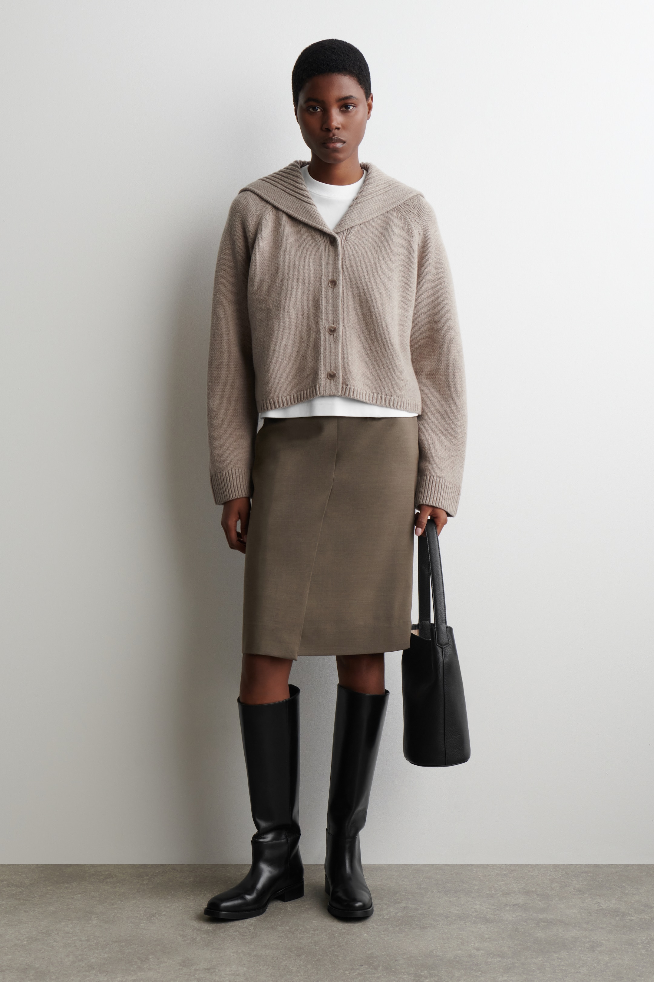 Visualizza immagine più grande: Un cardigan beige lavorato a maglia è indossato, rivolto in avanti, con un colletto a scialle a coste e una chiusura frontale con bottoni marroni. Ha maniche lunghe con polsini a coste e un orlo a coste, ed è indossato sopra una t-shirt bianca.