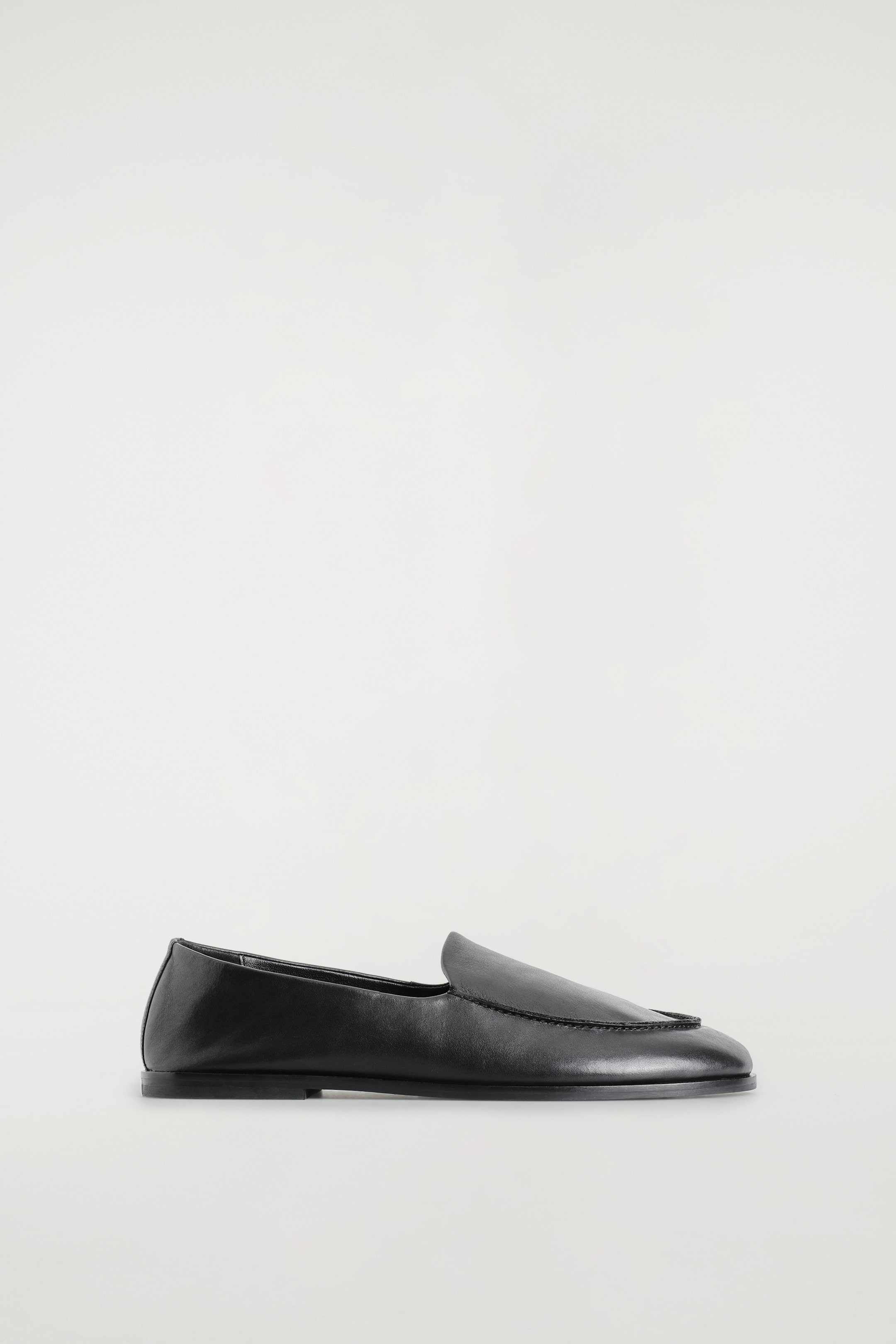 Visualizza immagine più grande: I classici mocassini in pelle presentano un design elegante slip-on in una vista di profilo laterale, con una tomaia in pelle nera liscia. La scarpa mostra una suola piatta, una punta arrotondata e sottili dettagli cuciti a grembiule attorno al dorso del piede.
