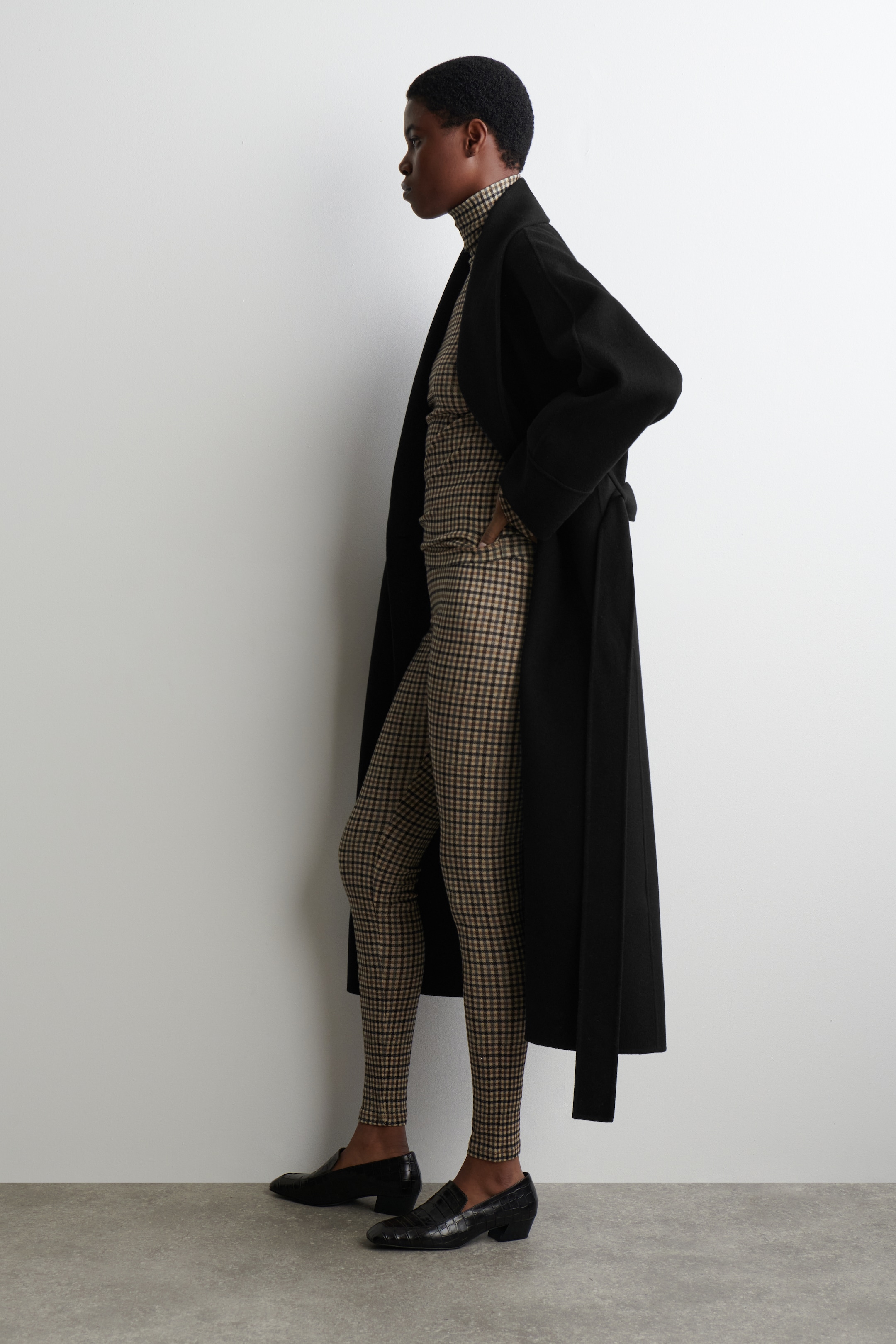 Visualizza immagine più grande: I leggings in lana merino a vestibilità slim, caratterizzati da un motivo a quadri beige e marrone scuro, sono indossati dalla modella con un dolcevita coordinato e un lungo cappotto nero.