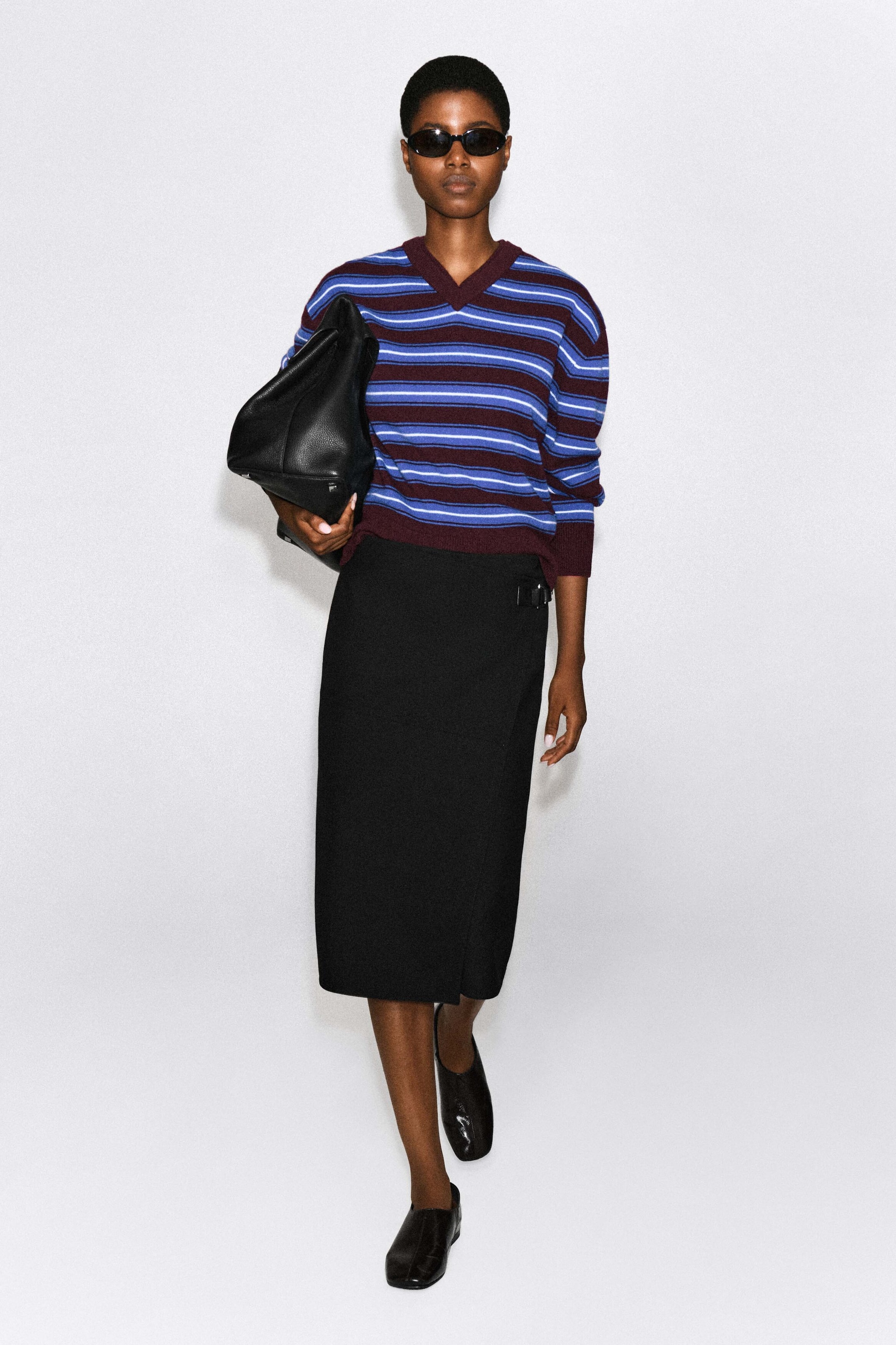 A Merino wool layered V-neck sweater, featuring horizontal stripes in shades of blue and burgundy with a double-layered ribbed collar, is worn by the model. The sweater is styled with a black midi skirt and a large black leather bag tucked under the arm. A Merino wool layered V-neck sweater, featuring horizontal stripes in shades of blue and burgundy with a double-layered ribbed collar, is worn by the model. The sweater is styled with a black midi skirt and a large black leather bag tucked under the arm.