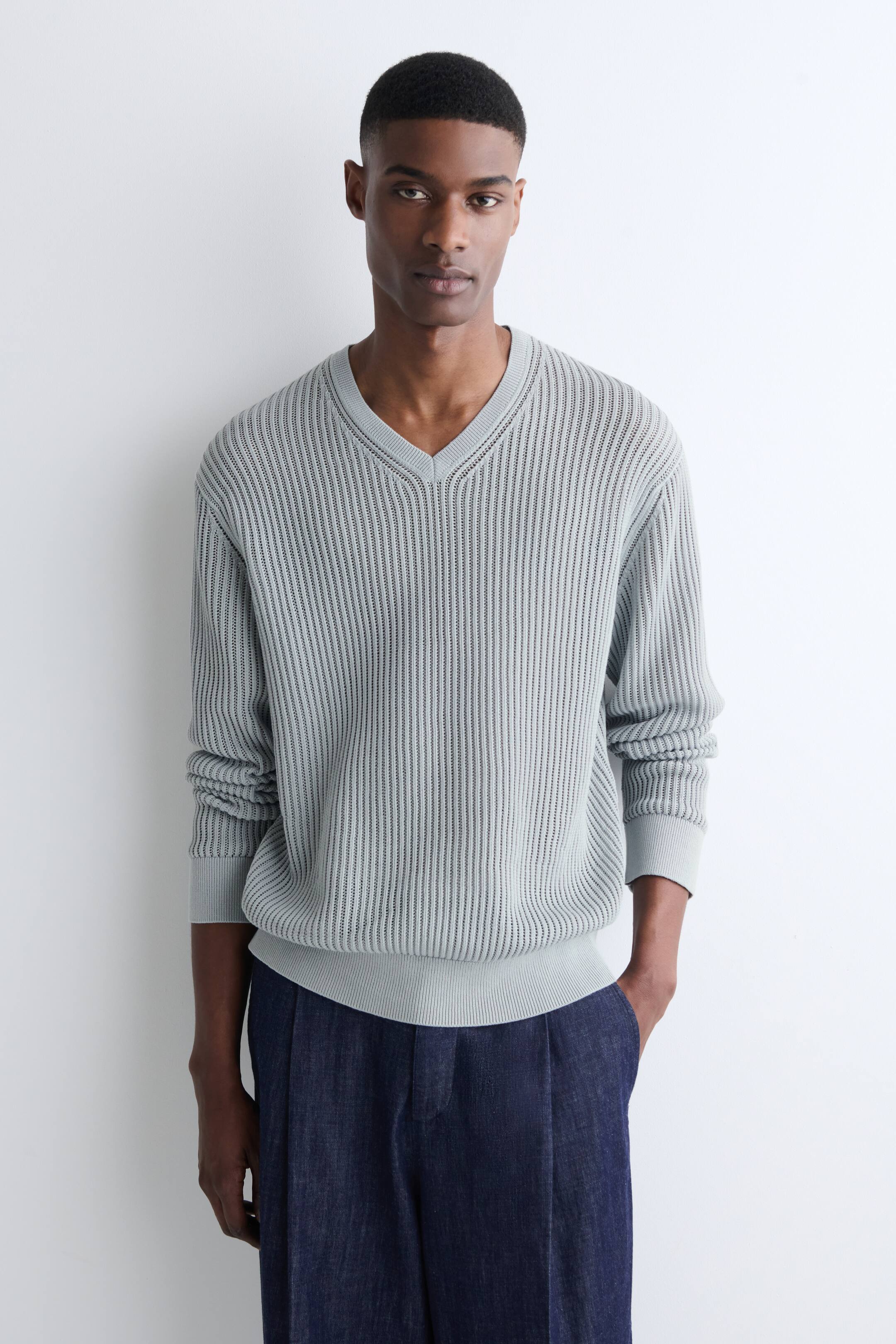 Visa större bild: En ljusblå V-ringad tröja bärs, med ett öppet vertikalt ribbstickat mönster över hela kroppen och långa ärmar. Den har en ribbad V-ringning, ribbade muddar och en ribbad fåll, och sitter i en avslappnad stil.