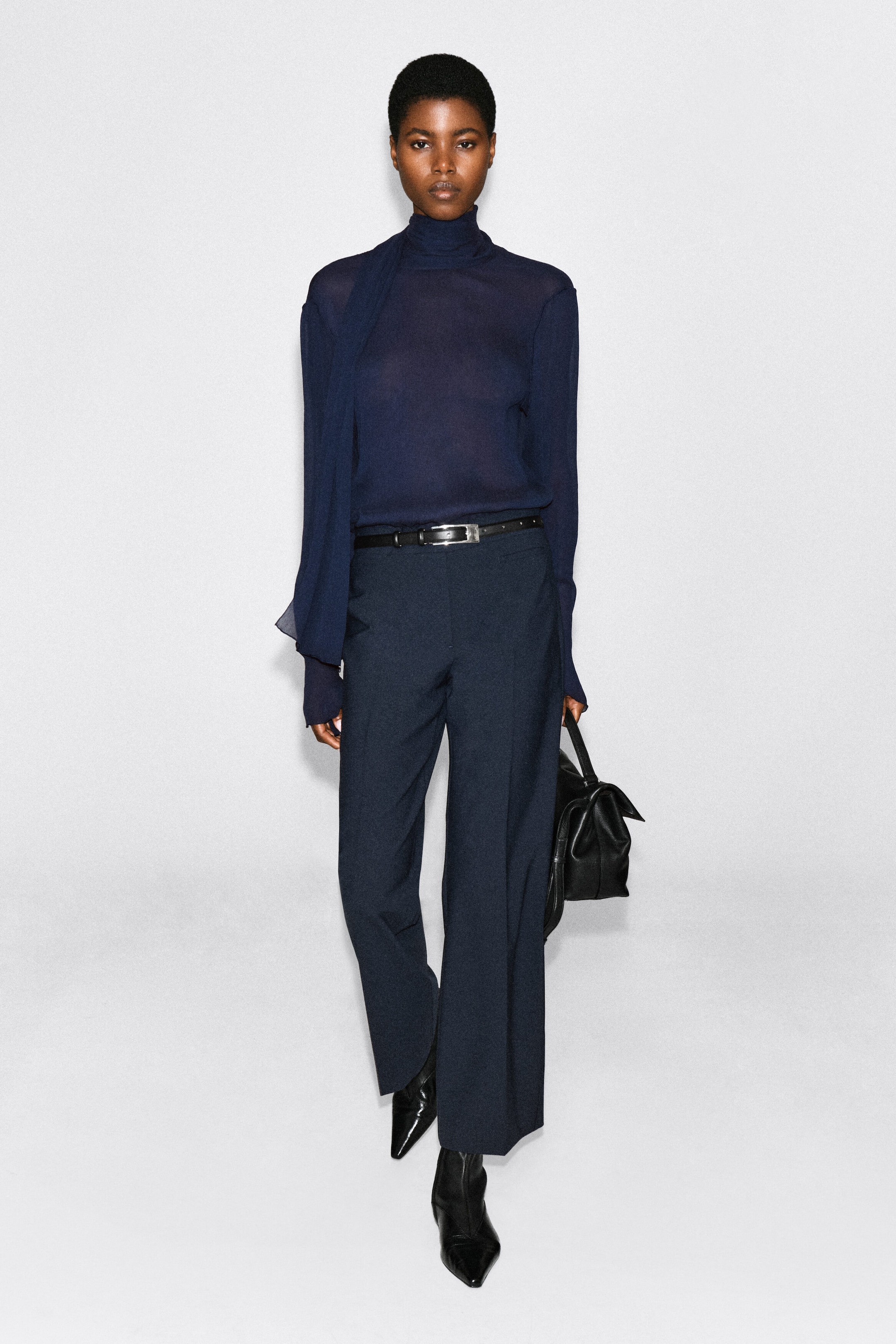 A semi-sheer, navy blue long-sleeved top featuring a high neck and a matching draped scarf is being worn by the model. The top is tucked into dark, wide-leg trousers styled with a black belt. A semi-sheer, navy blue long-sleeved top featuring a high neck and a matching draped scarf is being worn by the model. The top is tucked into dark, wide-leg trousers styled with a black belt.