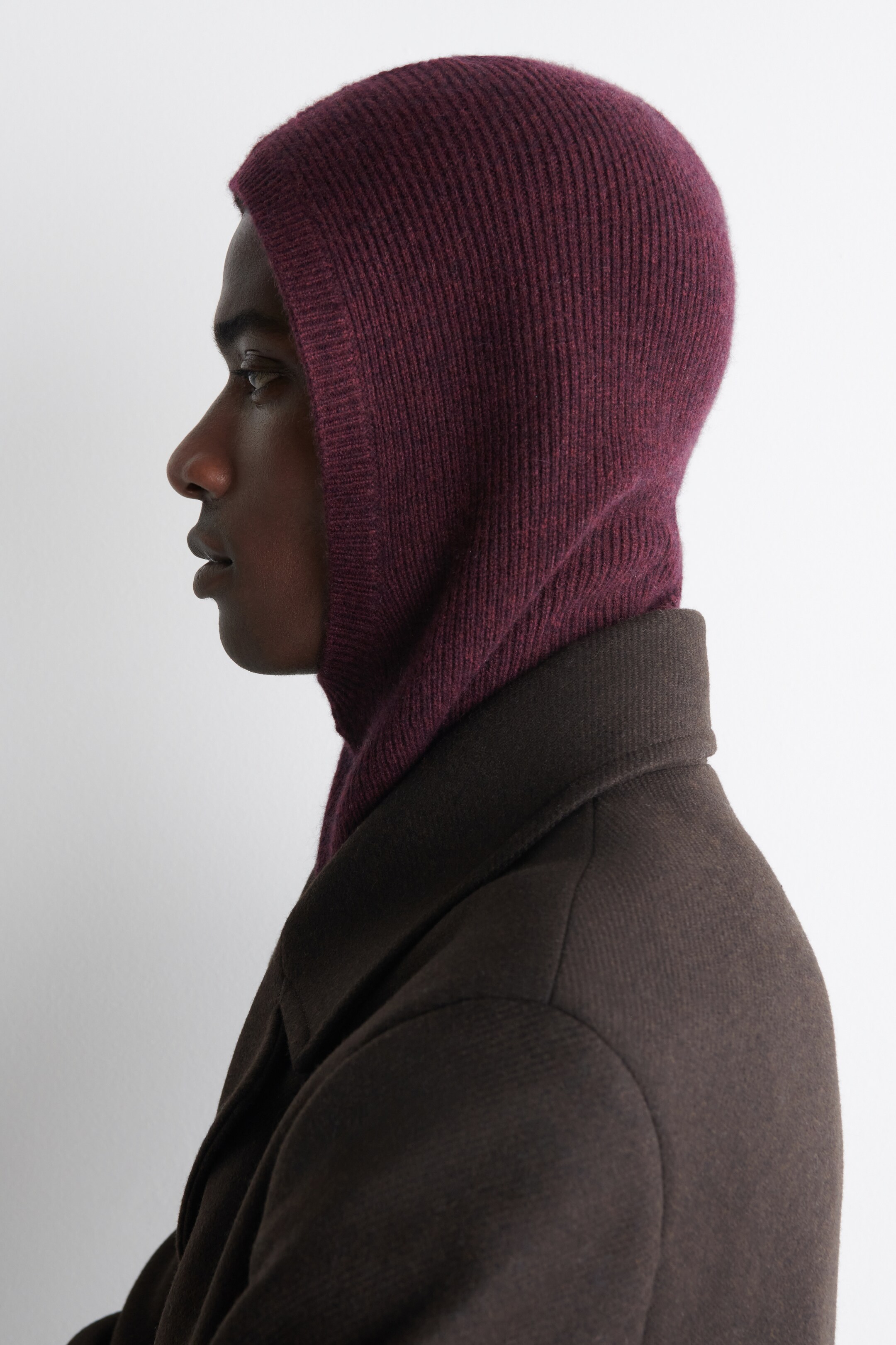 View larger image: A burgundy ribbed knit balaclava is covering the head and neck, fitting snugly. It is being worn layered under the dark brown collar of a coat, seen from a side profile.