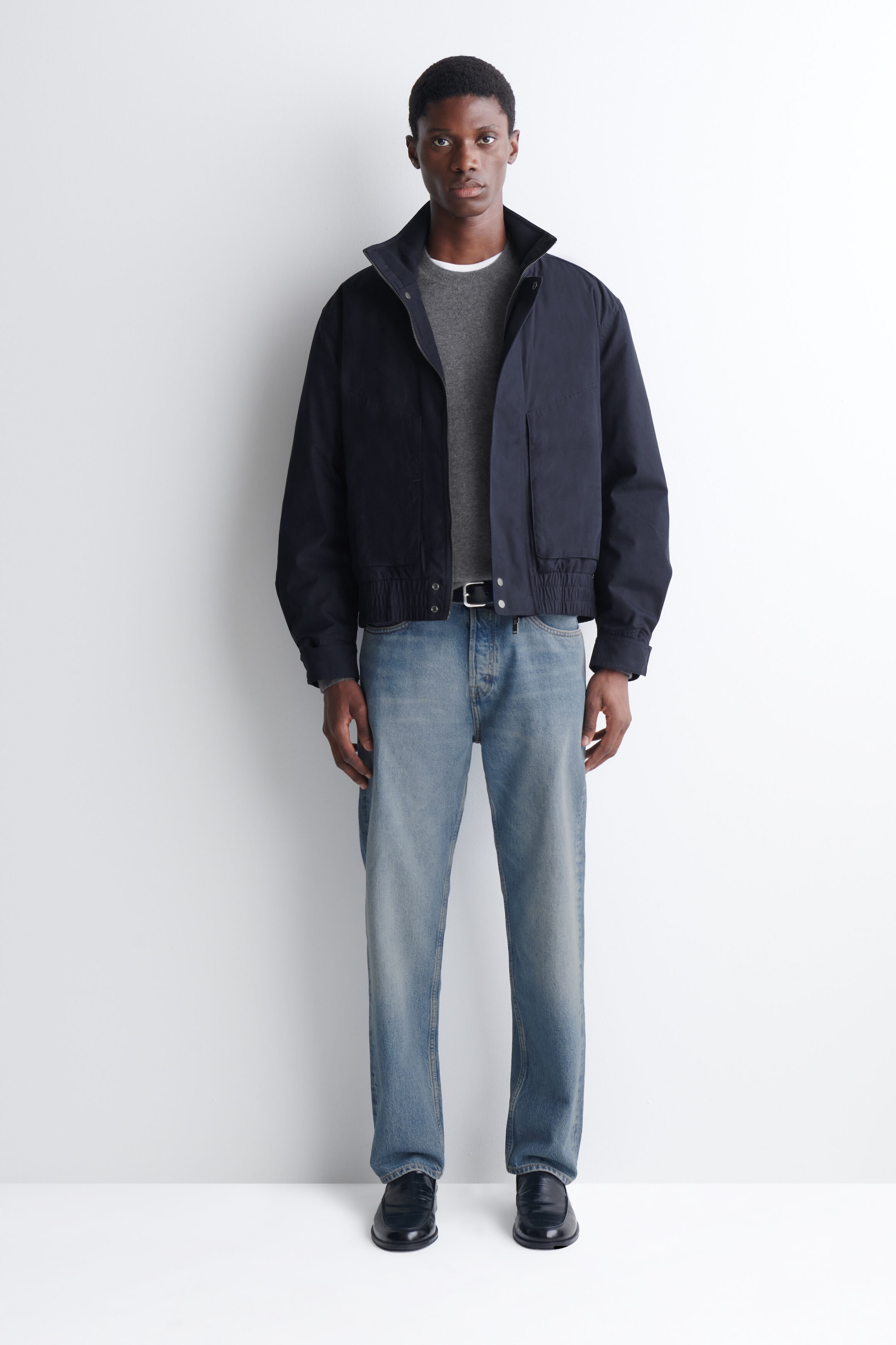 Une veste bomber bleu marine à col cheminée matelassé est portée ouverte par le mannequin, mettant en valeur son col montant, sa fermeture éclair à l’avant et son ourlet élastiqué. La veste est superposée à un chandail à col rond gris et agencée avec un jean droit bleu pâle.