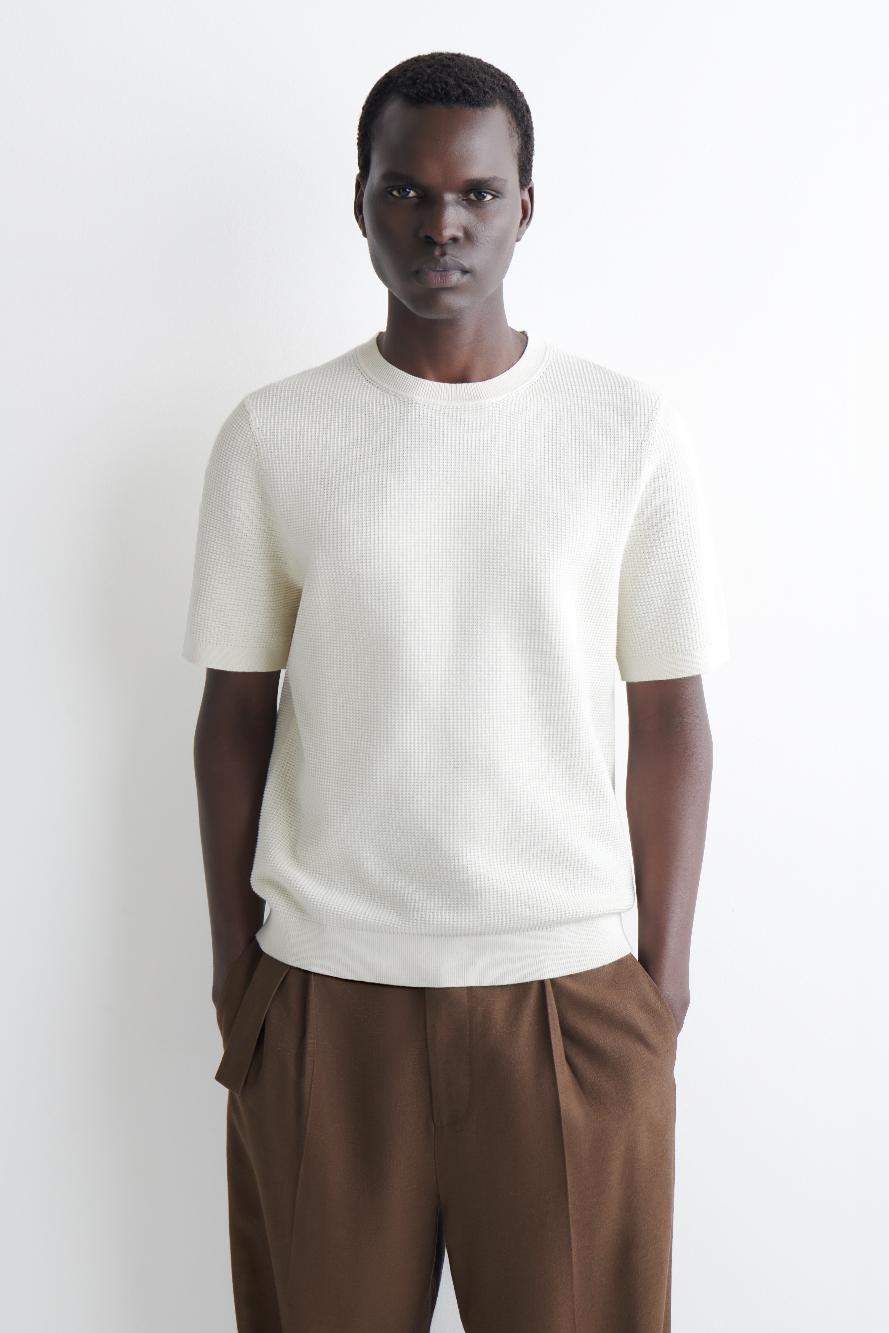 Una T-shirt bianco sporco è presentata dal FRONTE, mettendo in evidenza la sua texture a micro waffle. Presenta un collo a girocollo a coste, maniche corte con polsini a coste e un orlo a coste.