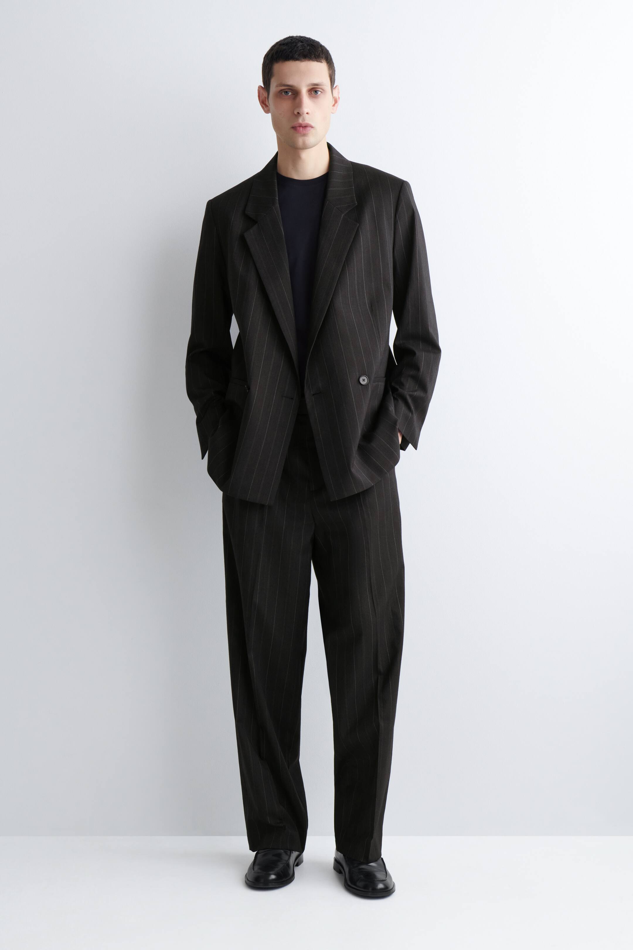 Преглед на по-голямо изображение: Тъмен костюм с фини райета е представен от преден изглед, включващ двуредно сако с назъбени ревери, закопчано с едно копче на талията и два джоба с капаци. Сакото е облечено върху черна тениска с кръгло деколте, а съответстващите широки панталони завършват ансамбъла.