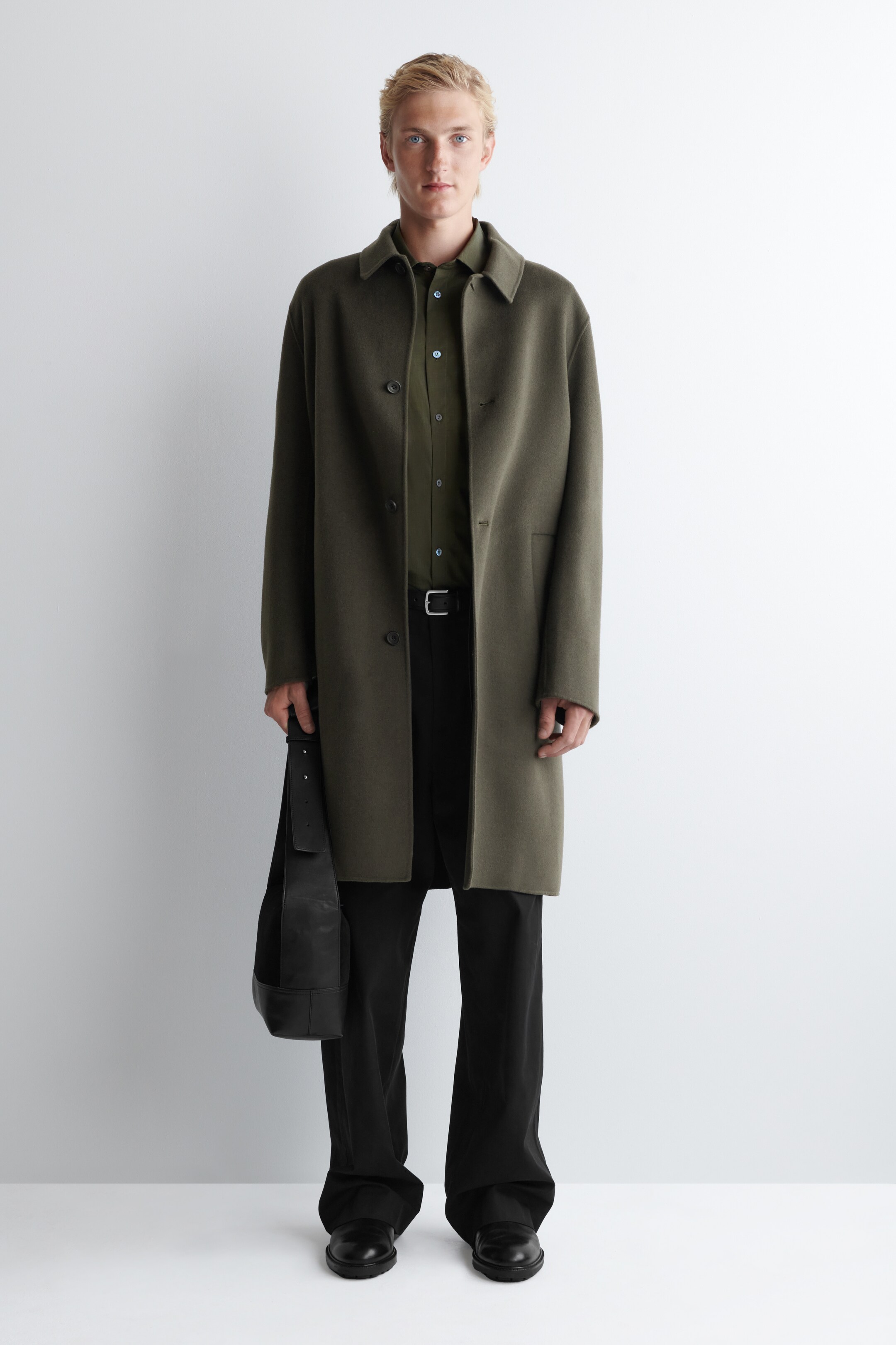 Ver imagen más grande: Un abrigo verde oliva de cara frontal se lleva puesto, con un cuello en punta, tres botones visibles en la parte delantera y dos bolsillos de ojal laterales. Está superpuesto sobre una camisa verde oliva más oscura y pantalones negros, con un bolso de hombro de cuero negro sostenido al costado.