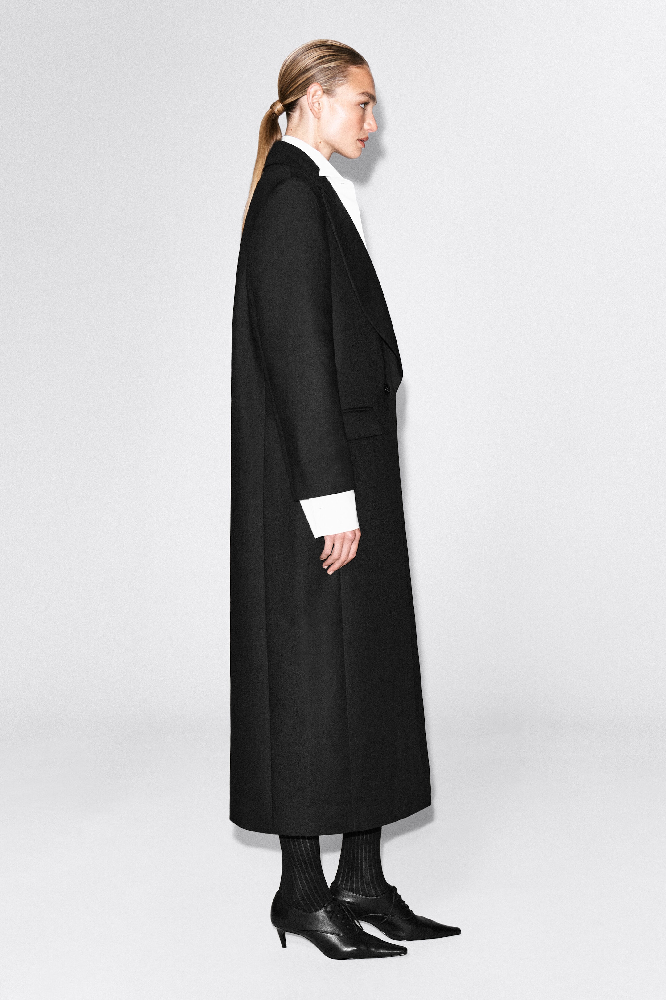 The model is wearing a long black wool coat, featuring a relaxed silhouette, structured lapels and a single-breasted front. The coat is layered over a white shirt with a visible collar and cuffs, and is styled with black ribbed tights and pointed-toe lace-up heels.
