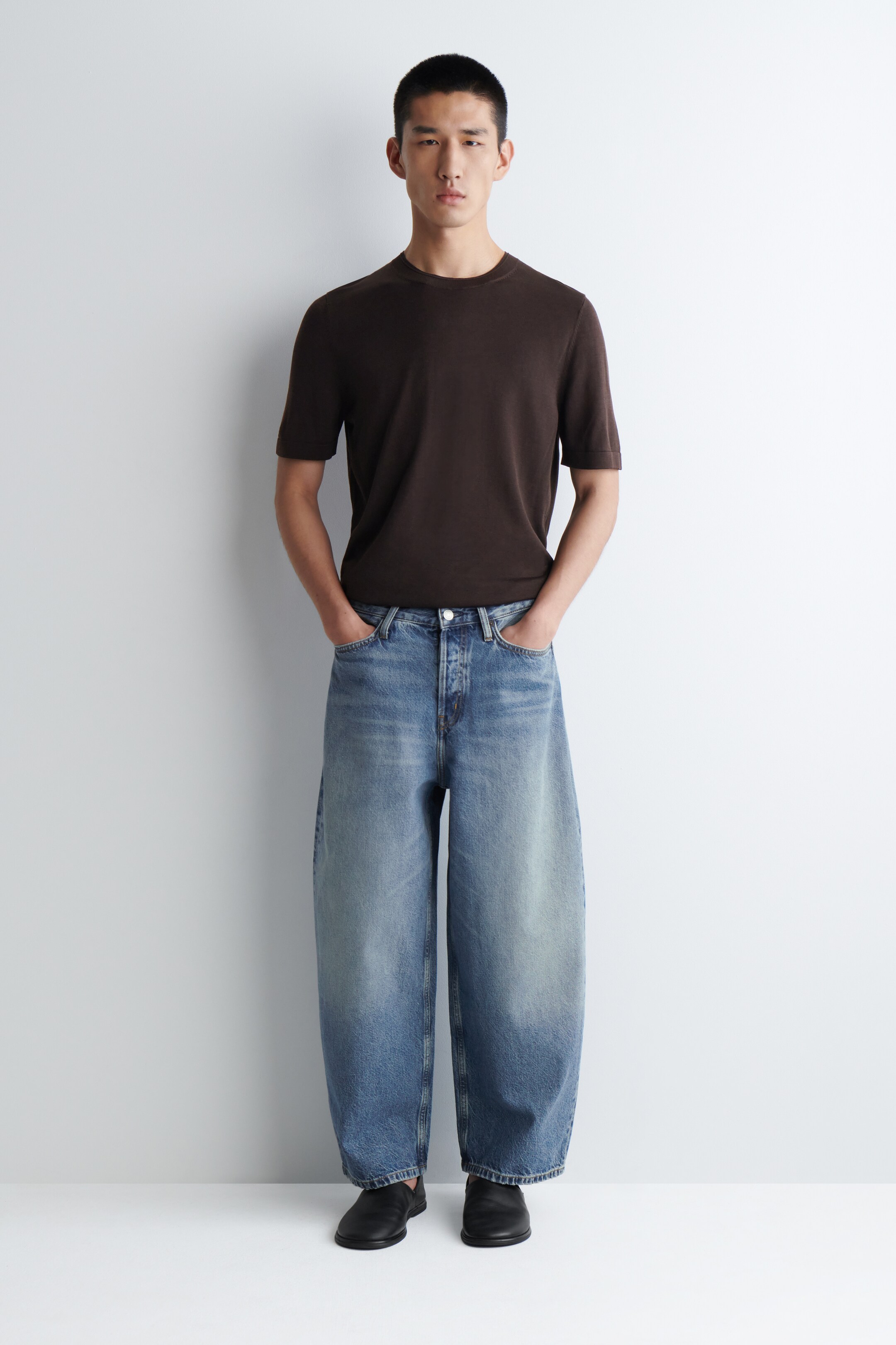 Agrandir l'image: Un t-shirt en tricot marron foncé à manches courtes avec un col rond est porté, assorti à un jean en denim bleu clair à jambes larges. Le jean présente une braguette à boutons et un délavage décoloré. Des chaussures noires à enfiler complètent le look.