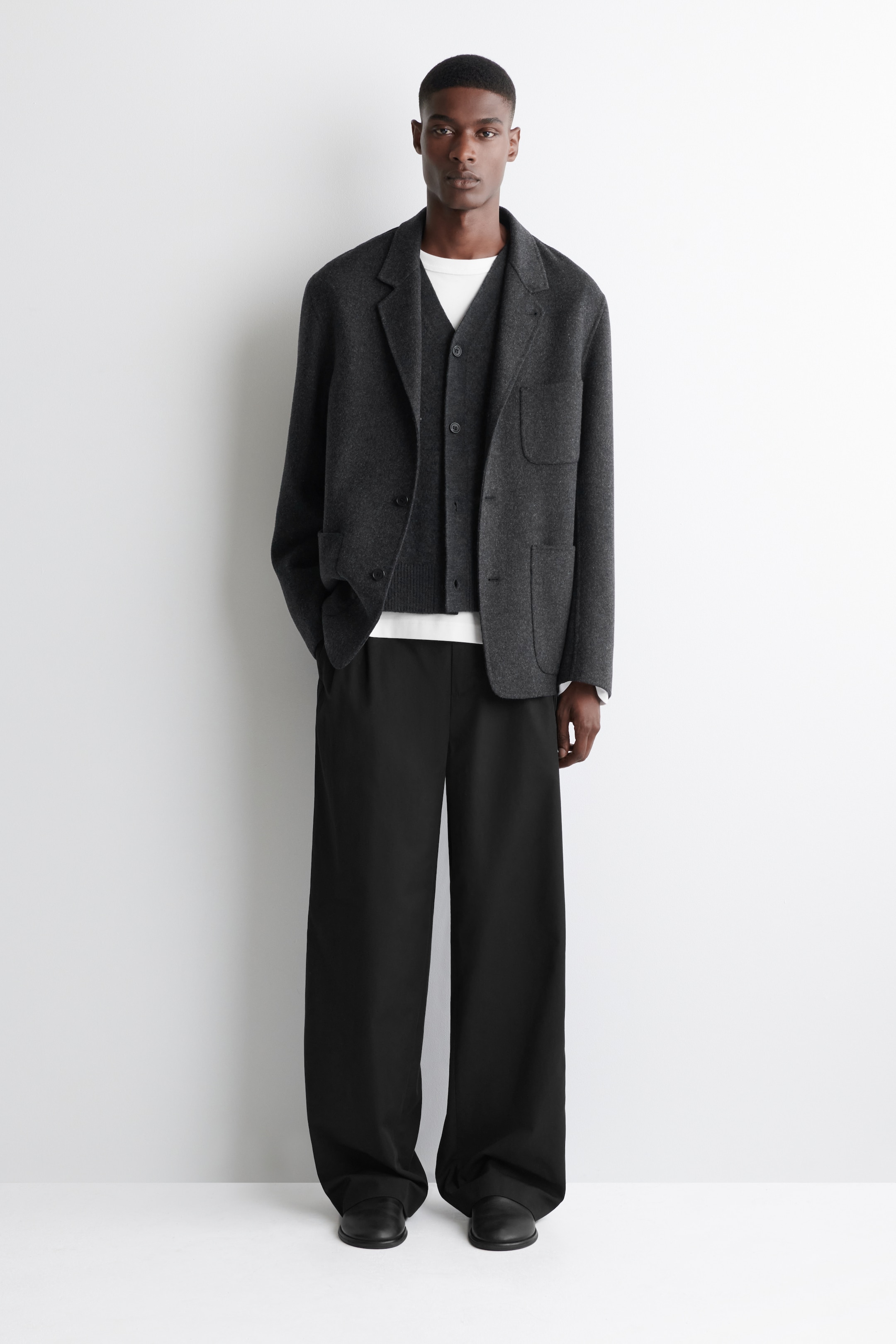 View larger image: A men's ensemble is featuring a dark gray, relaxed-fit blazer with notch lapels and two patch pockets, layered over a dark gray V-neck cardigan and a white crew-neck t-shirt. Black, wide-leg trousers and black slip-on shoes are completing the look, all facing straight on.