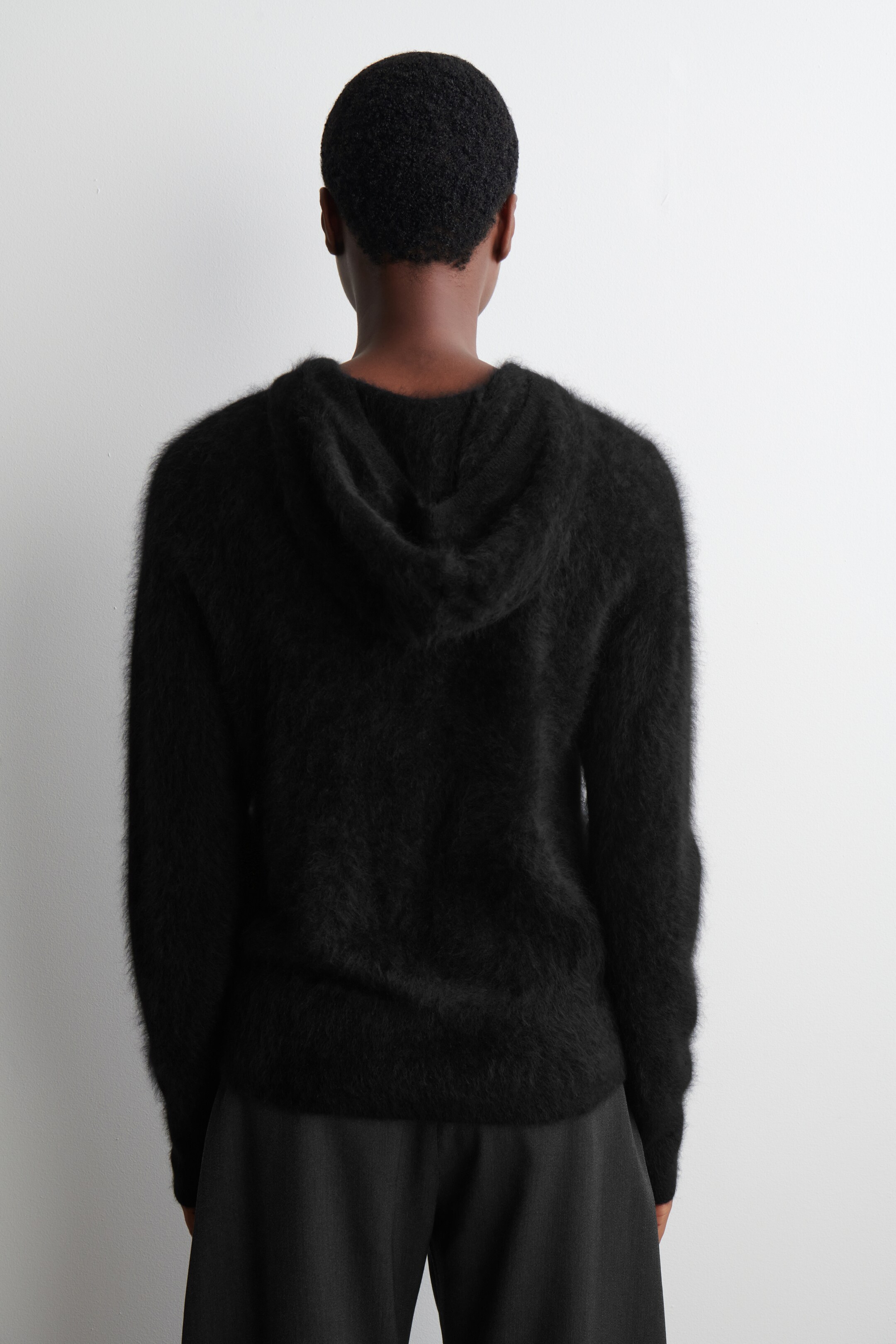 View larger image: A black fuzzy hooded sweater is being presented from the back, showcasing its relaxed fit and shaggy texture. The sweater is featuring a visible hood and long sleeves, and is being worn over dark trousers.
