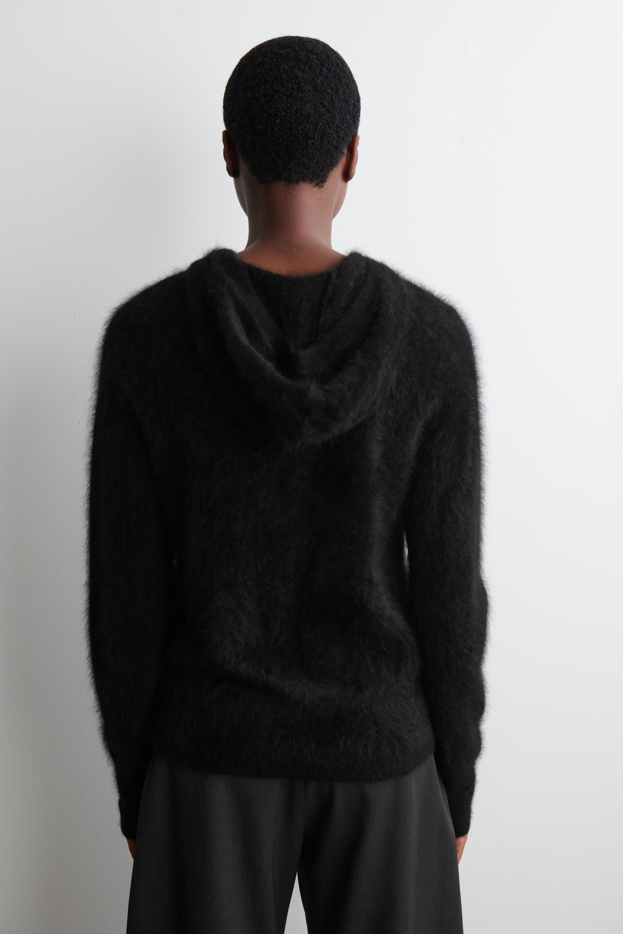 View larger image: A black fuzzy hooded sweater is being presented from the back, showcasing its relaxed fit and shaggy texture. The sweater is featuring a visible hood and long sleeves, and is being worn over dark trousers.