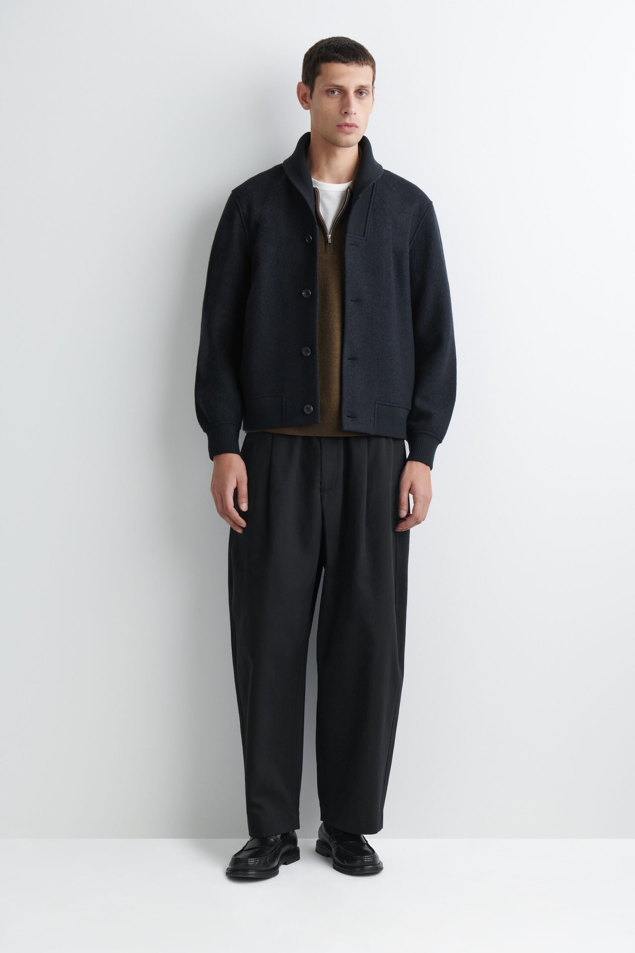 The navy boiled-wool shawl-collar jacket features a structured shawl collar, a button-front closure, and ribbed detailing at the cuffs and hem in a front-facing view. It is styled open over a brown half-zip sweater, highlighting its relaxed fit and heavy, textured fabric. The navy boiled-wool shawl-collar jacket features a structured shawl collar, a button-front closure, and ribbed detailing at the cuffs and hem in a front-facing view. It is styled open over a brown half-zip sweater, highlighting its relaxed fit and heavy, textured fabric.