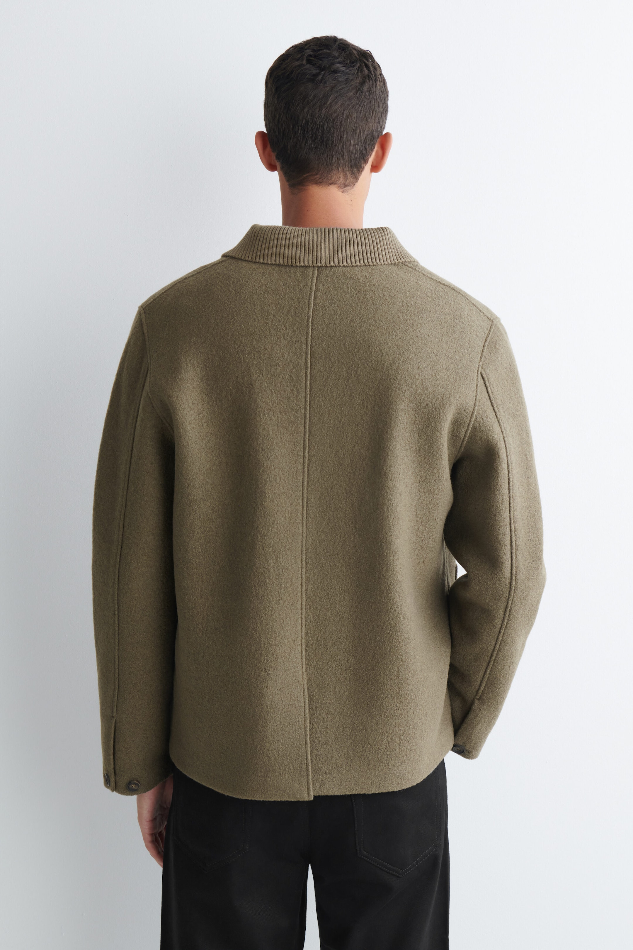 A ribbed-collar boiled-wool chore jacket in olive green is worn by the model, shown from the back. The jacket features a distinct ribbed-knit collar, a center back seam, and buttoned cuffs, and is styled with black trousers. A ribbed-collar boiled-wool chore jacket in olive green is worn by the model, shown from the back. The jacket features a distinct ribbed-knit collar, a center back seam, and buttoned cuffs, and is styled with black trousers.