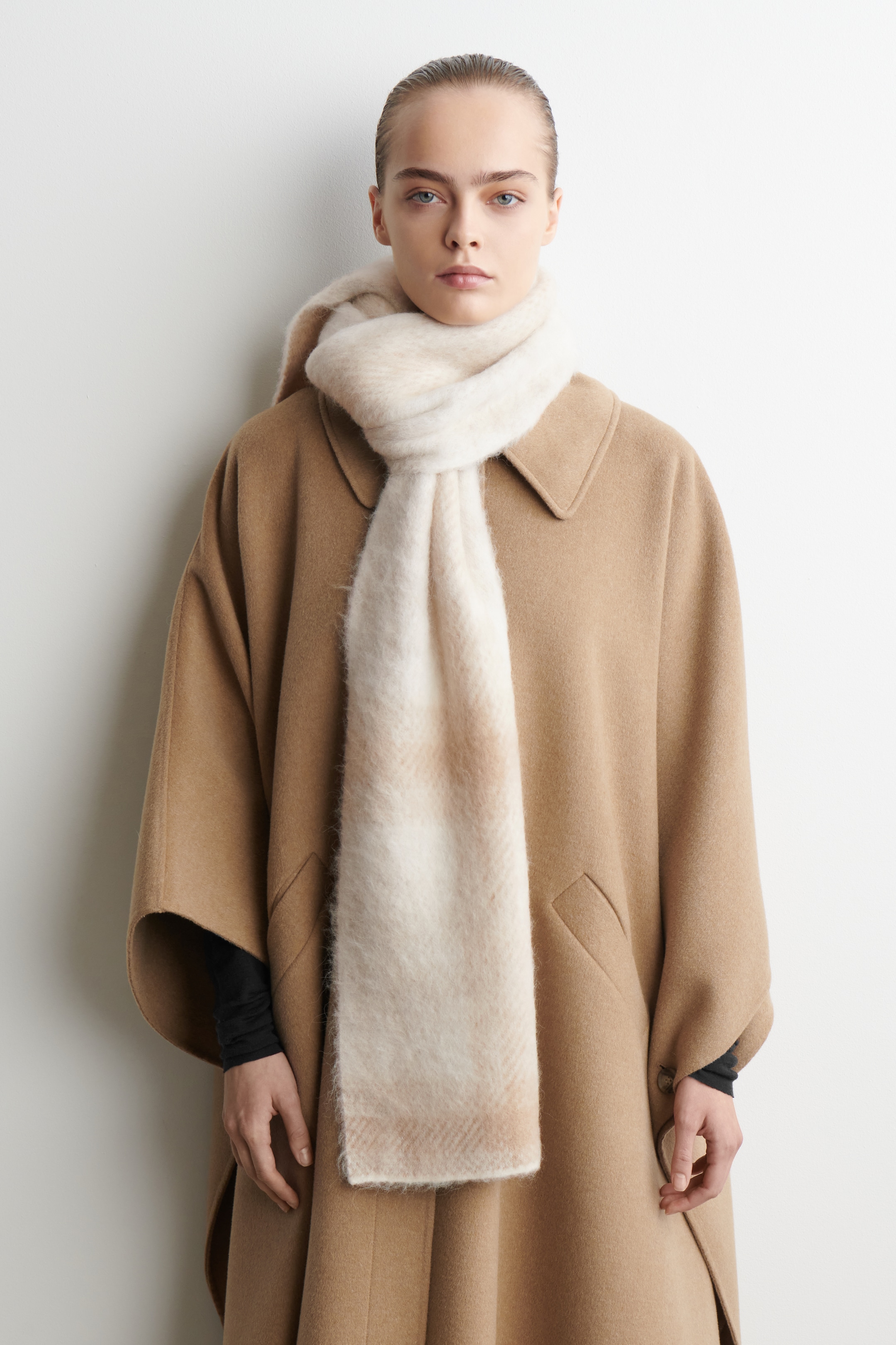 Agrandir l'image: Une écharpe brossée, ornée d'un motif à carreaux ombré subtil en crème et beige clair, est enroulée autour du cou du mannequin. L'écharpe est drapée sur un manteau de couleur camel, avec une extrémité pendante sur le devant.