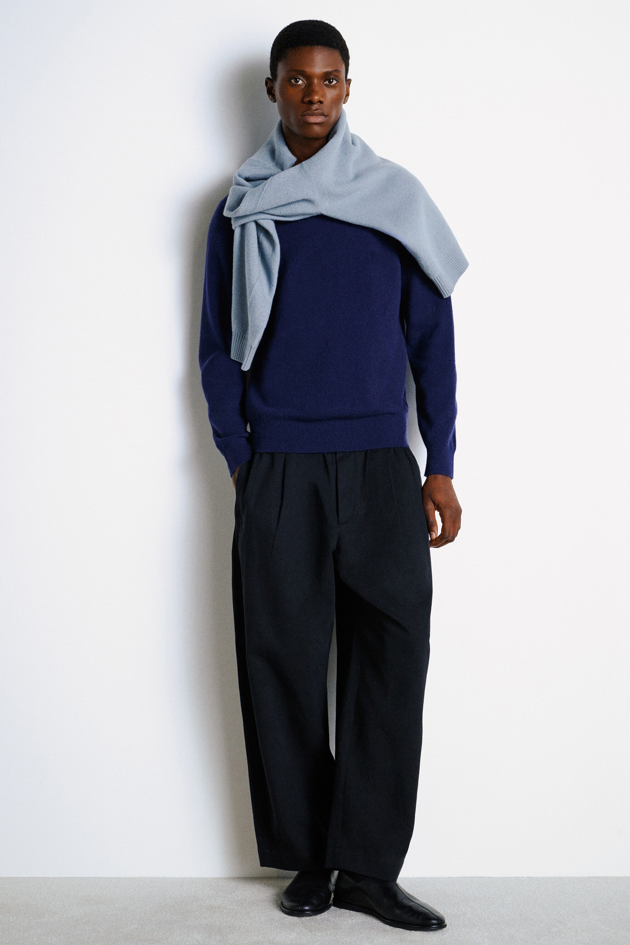Un pull en cachemire à col rond bleu foncé est présenté de face, avec une coupe régulière, des poignets côtelés et un ourlet côtelé. Un pull en tricot bleu pâle est drapé sur les épaules, porté avec un pantalon noir à jambe large.