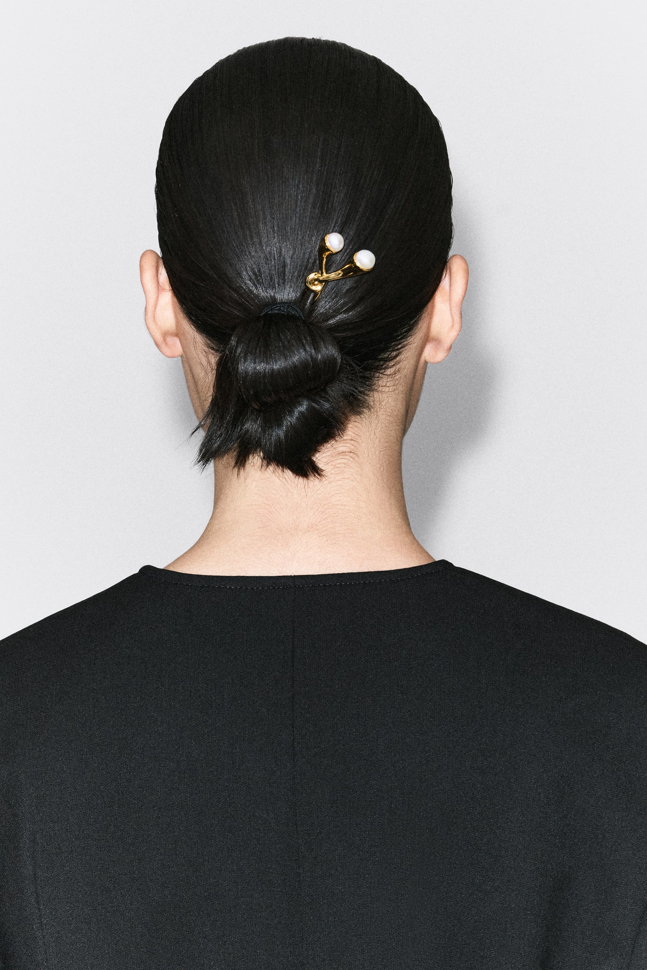 Agrandir l'image: Une épingle à cheveux dorée, présentant une forme sculptée de manière organique et ornée de deux perles blanches à son extrémité, est montrée de dos, maintenant un chignon bas plaqué et noué. L’épingle est positionnée en diagonale au-dessus du chignon, mettant en valeur son design torsadé et ses accents de perles.