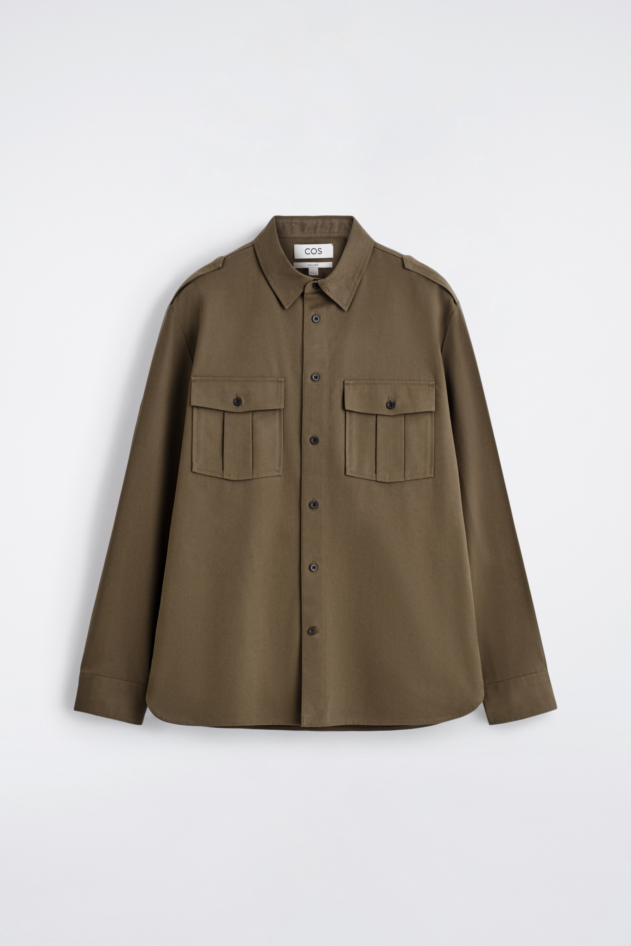 Ver imagen más grande: Una camisa verde oliva oscuro se presenta desde el frente, con un cuello en punta, un cierre completo de botones en la parte delantera con botones oscuros, y dos grandes bolsillos en el pecho con solapas abotonadas. Las charreteras son visibles en cada hombro, y la camisa está diseñada con mangas largas y un ajuste holgado.