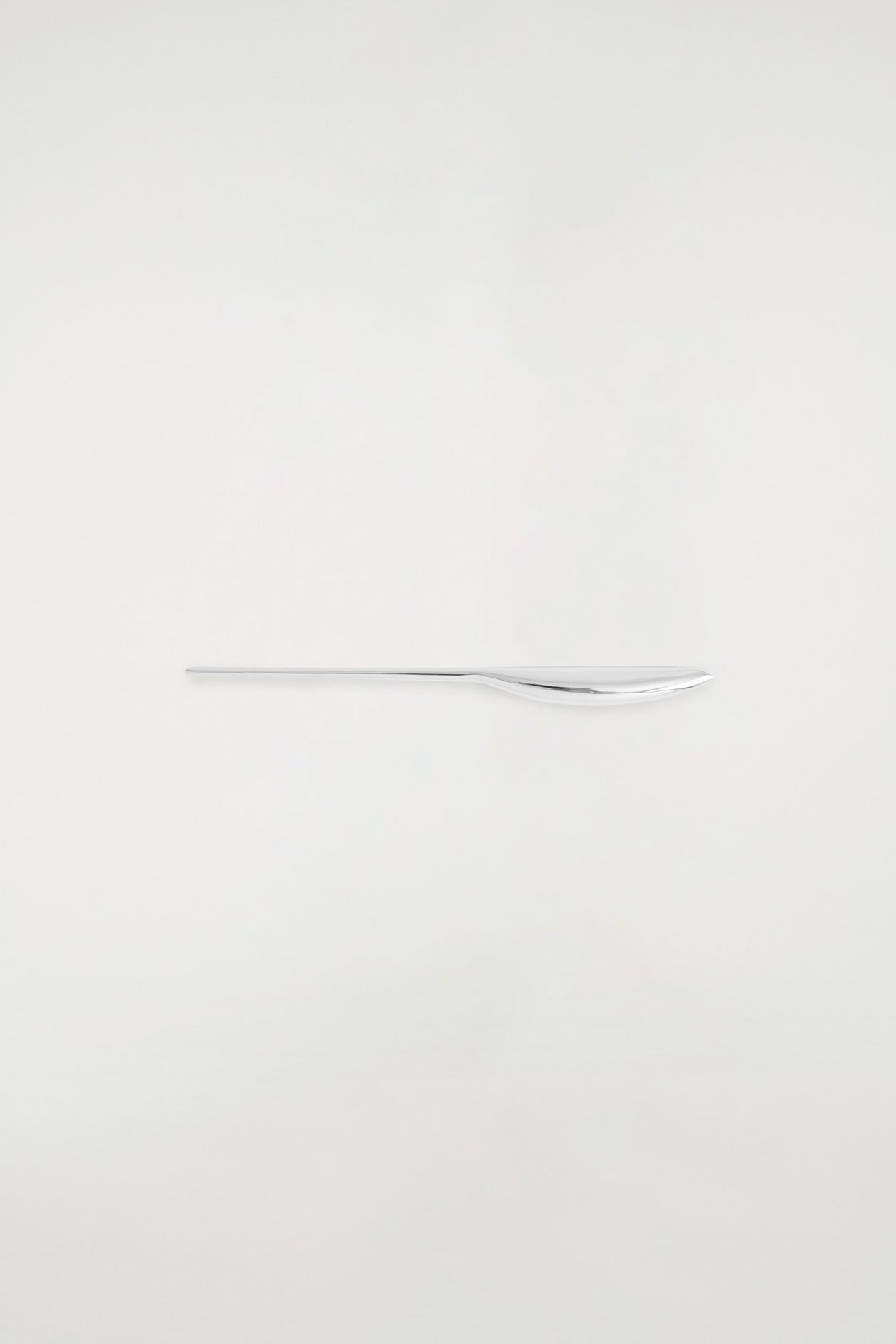 A polished silver-toned knife lies horizontally, featuring a minimalist design with a long, ultra-thin handle on the left and a smoothly curved, reflective blade on the right.