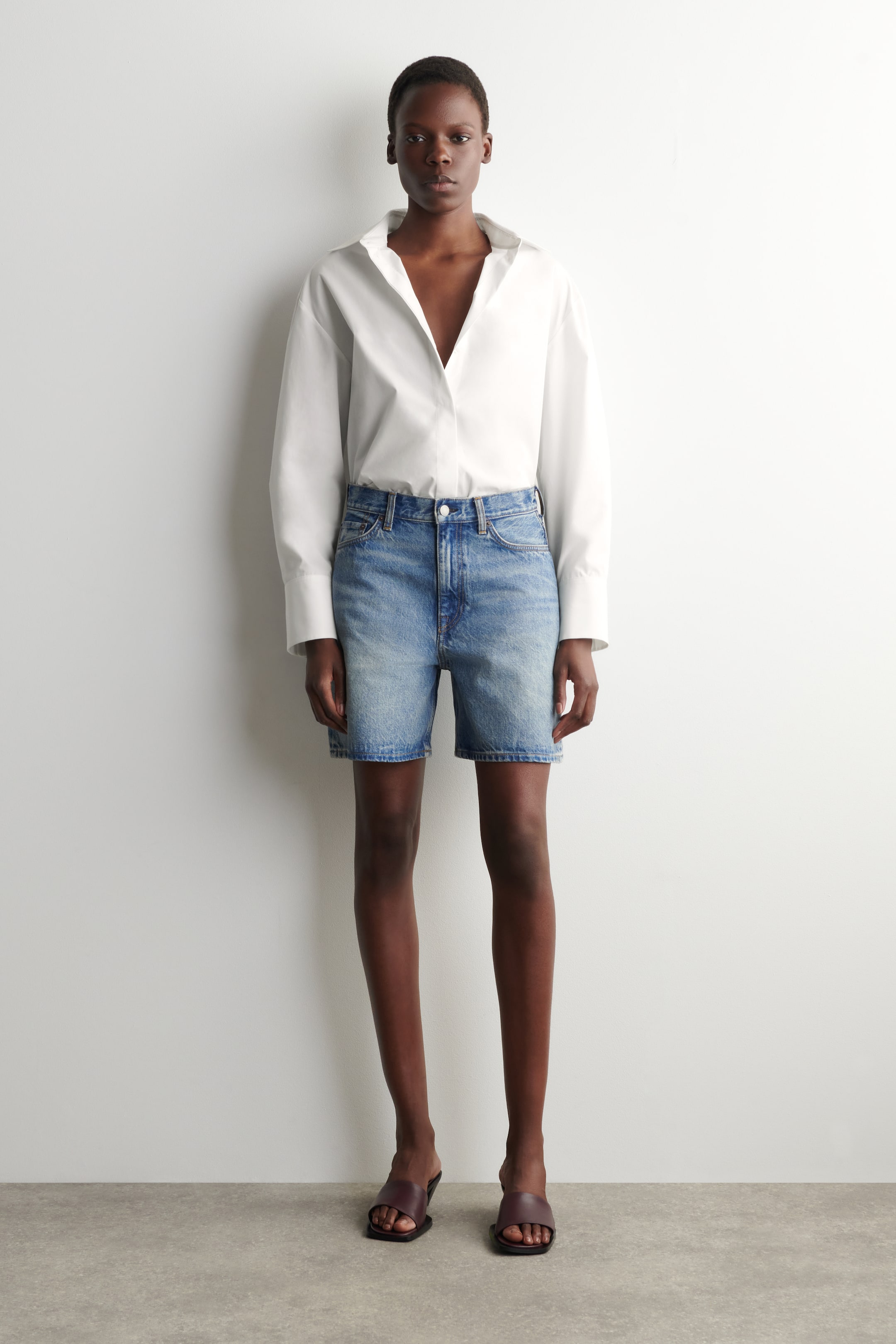 הצגת תמונה גדולה יותר: חולצה לבנה עם שרוולים ארוכים, בעלת צווארון פתוח ושרוולים רחבים, נלבשת תחובה לתוך מכנסי ג'ינס קצרים בצבע כחול בהיר. המכנסיים בעלי מותן גבוהה מציגים שטיפה דהויה ומגיעים עד אמצע הירך, בעוד סנדלים כהים בצבע חום נלבשים.