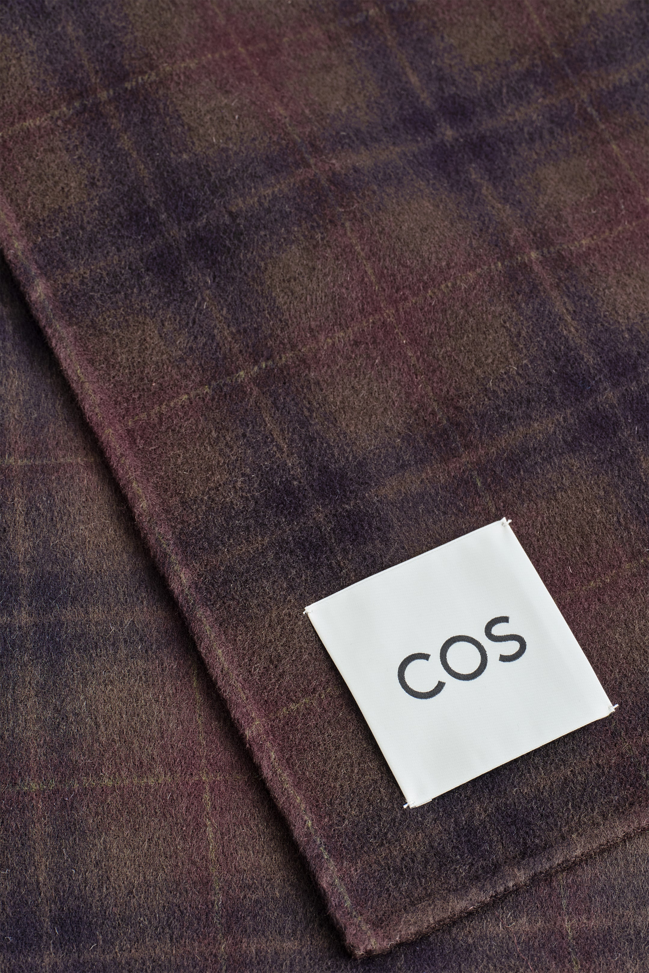 Agrandir l'image: Une écharpe en tartan affiche un motif à carreaux dans des nuances de bordeaux et de brun foncé. Un écusson rectangulaire blanc avec 'COS' en lettres noires est cousu à une extrémité, face à l'AVANT.