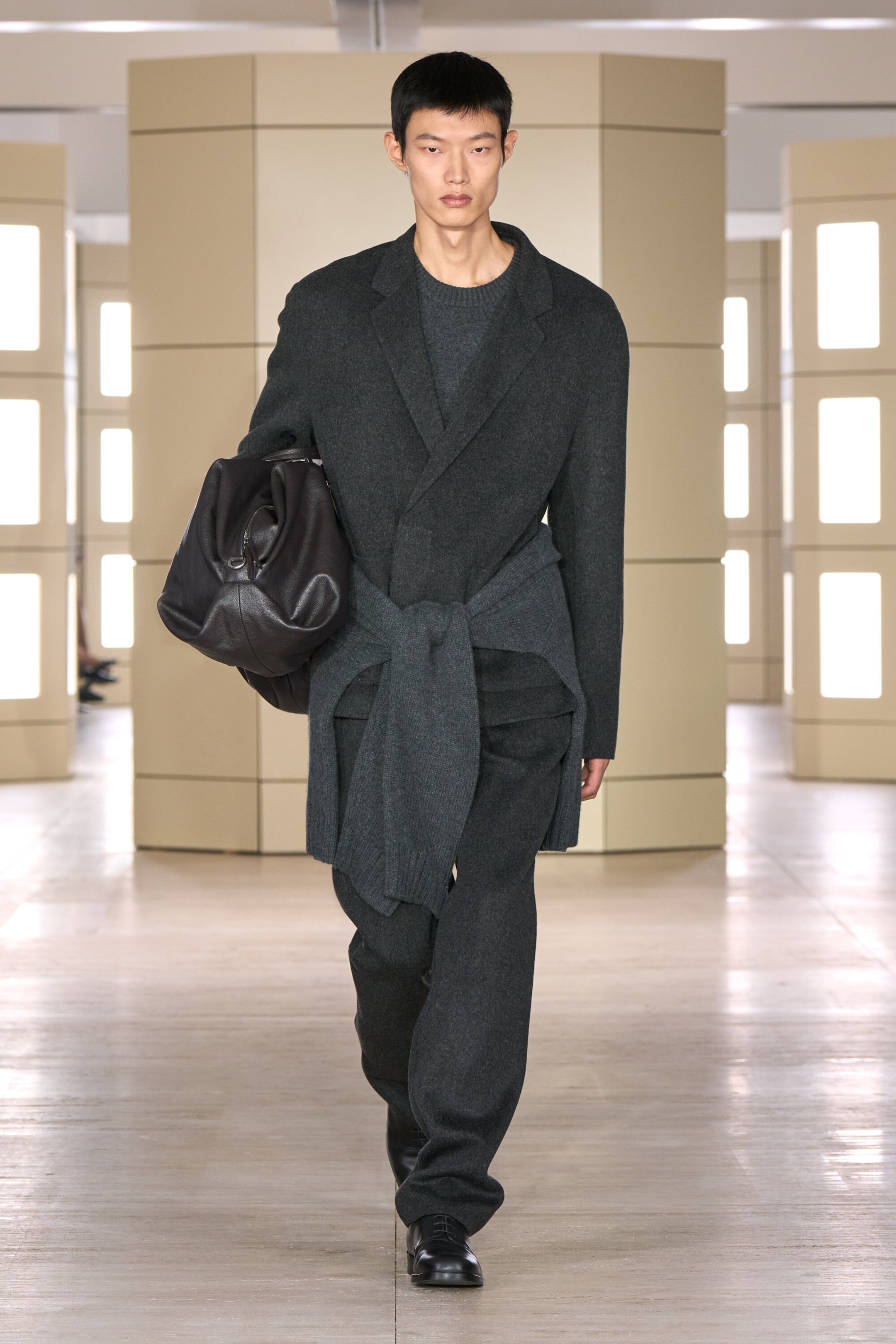 Presenting a front view, the model is wearing a dark grey, unstructured double-faced wool-blend blazer and matching straight-leg trousers. The relaxed-fit suit is styled with a grey crew-neck jumper underneath, a second grey jumper tied around the waist, and a large black leather bag held under the arm. Presenting a front view, the model is wearing a dark grey, unstructured double-faced wool-blend blazer and matching straight-leg trousers. The relaxed-fit suit is styled with a grey crew-neck jumper underneath, a second grey jumper tied around the waist, and a large black leather bag held under the arm.