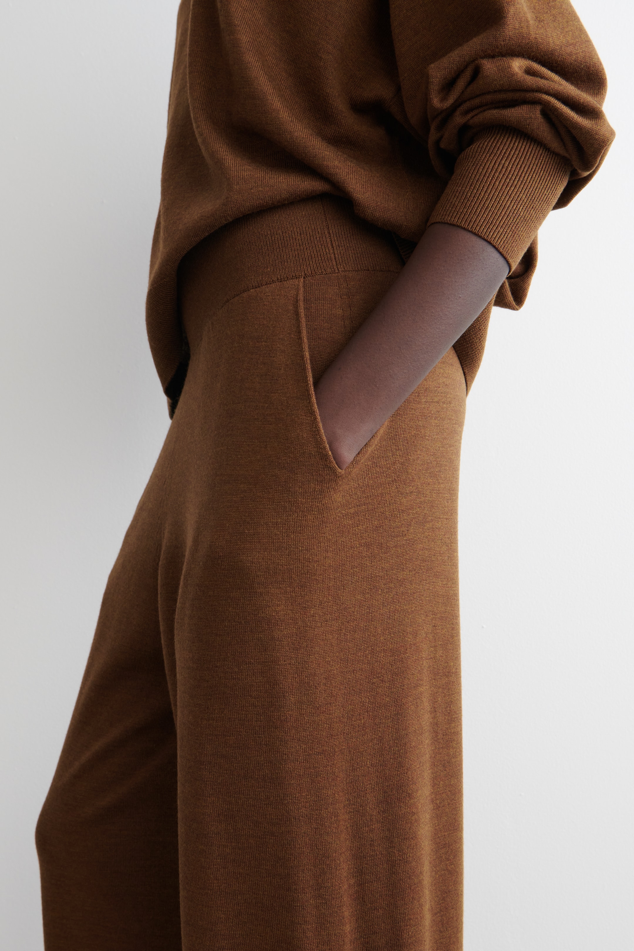 Visualizza immagine più grande: Un top marrone lavorato a maglia e pantaloni coordinati vengono indossati, visti di lato. Il top presenta maniche voluminose con polsini a coste e un orlo a coste, mentre i pantaloni sono progettati con una larga fascia in vita a coste e tasche laterali funzionali, in una delle quali sta entrando un braccio.