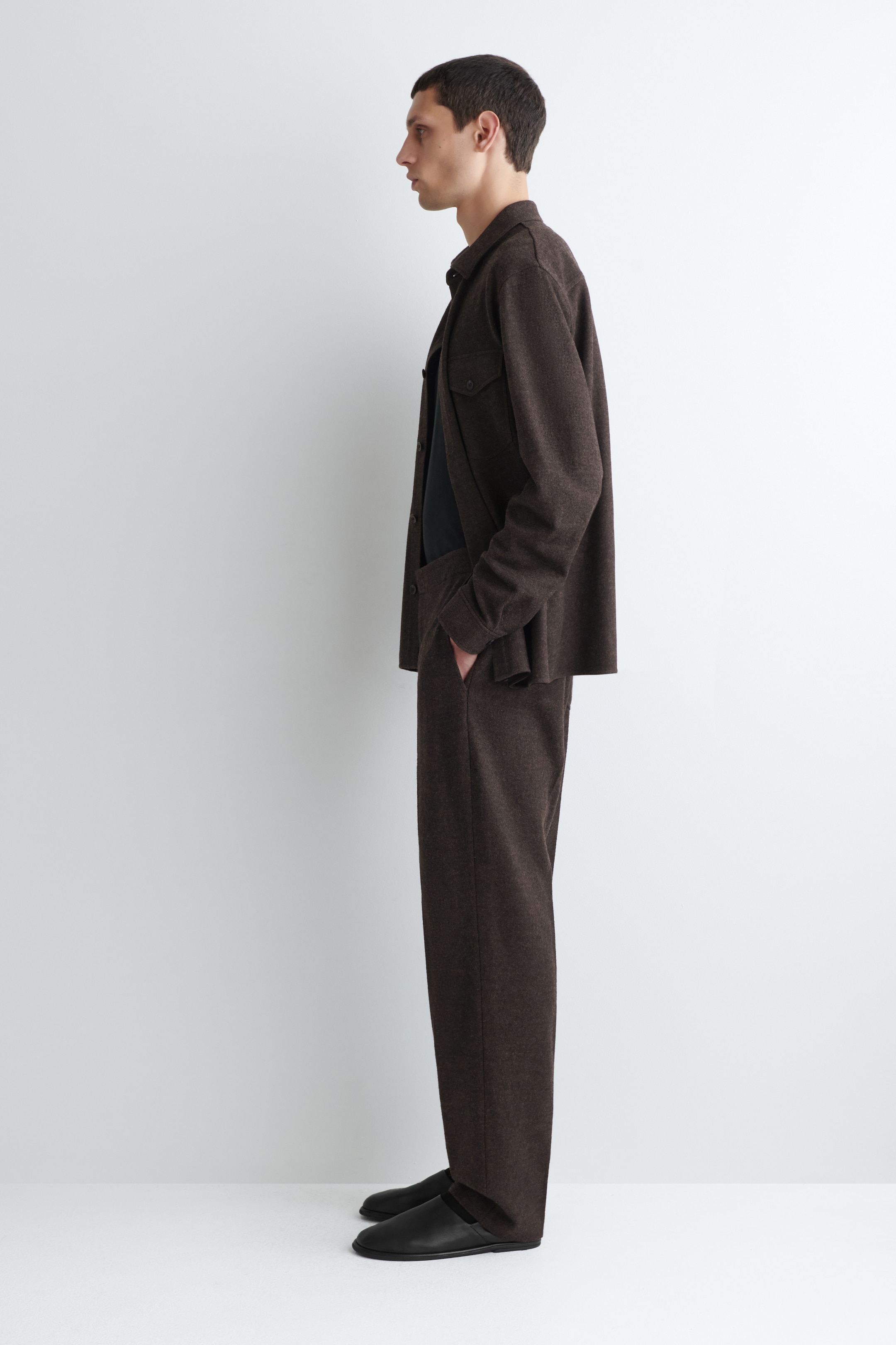 Visualizza immagine più grande: Una shacket marrone scuro e pantaloni coordinati a vestibilità rilassata vengono presentati di profilo. La shacket presenta un colletto, una chiusura a bottoni e due tasche sul petto con patte, ed è indossata aperta su una maglietta scura. I pantaloni a gamba dritta completano l'ensemble, che è abbinato a scarpe nere senza lacci.