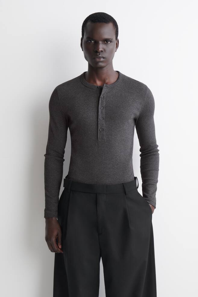 Den slanke langÃ¦rmede henley T-shirt i ribbet bomuld vises forfra og fremhÃ¦ver et mÃ¸rkegrÃ¥t ribbet stof, en rund halsudskÃ¦ring og en knappet stolpelukning foran. Skjorten sidder tÃ¦t til kroppen og er stukket ned i sorte bukser for at fremhÃ¦ve det slim-fit skÃ¥ret design. - 1