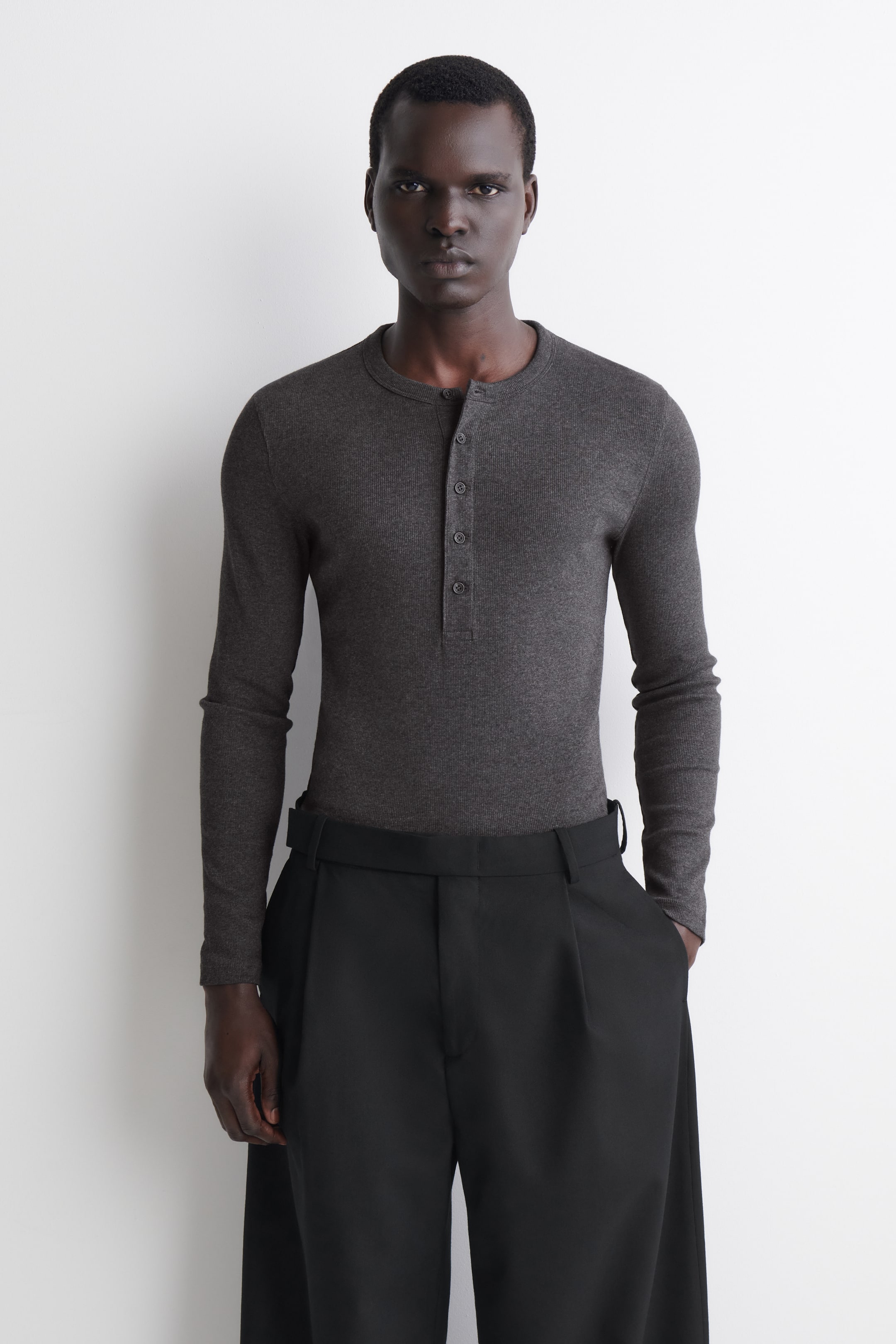 Grotere afbeelding bekijken: Het slim fit, geribde katoenen henley T-shirt met lange mouwen wordt van voren getoond en laat een donkergrijze geribde stof, een ronde hals en een geknoopte knooplijst vooraan zien. Het shirt sluit nauw aan op het lichaam en is ingestopt in een zwarte broek om het slanke model extra te benadrukken.