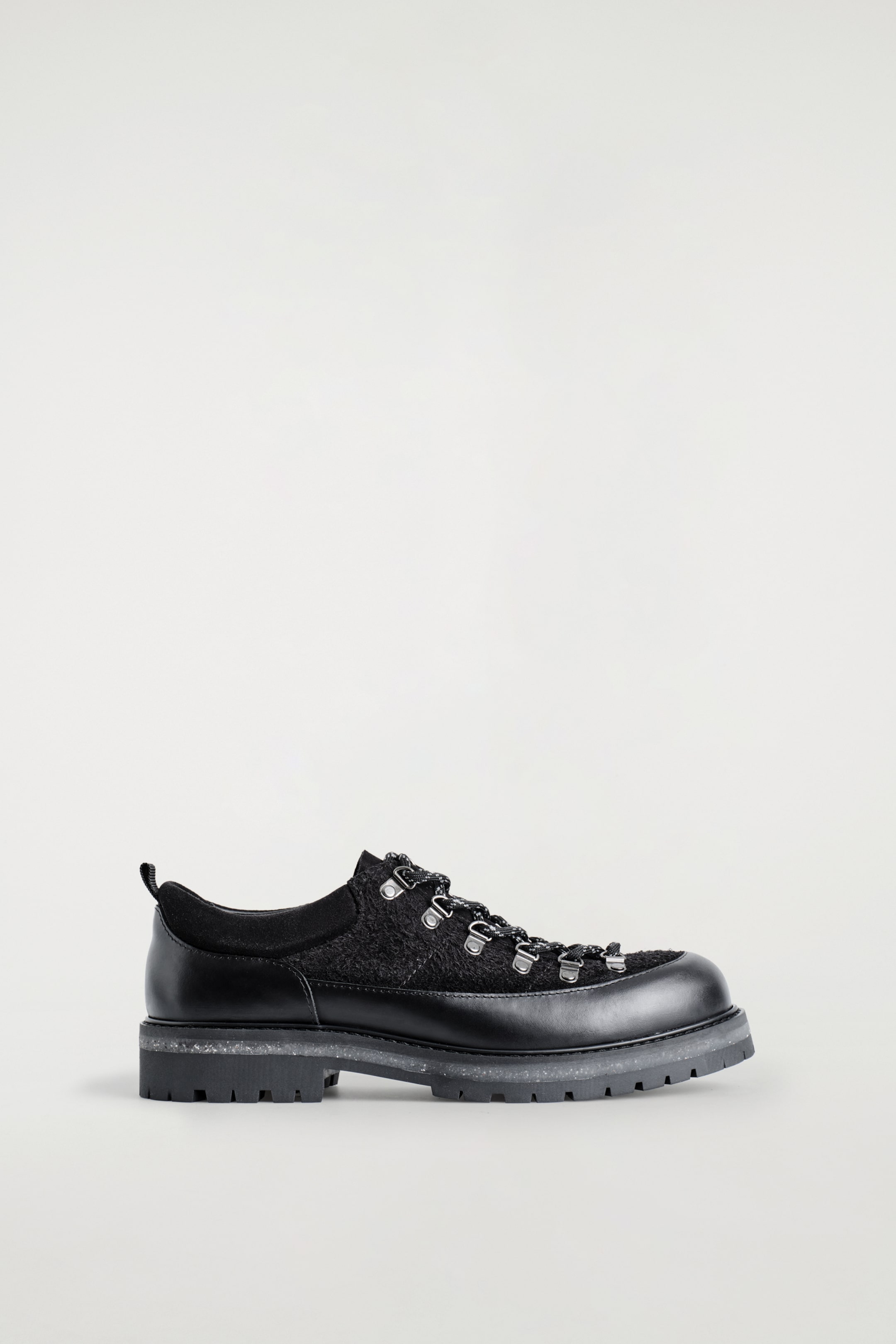 Ver imagen más grande: Se presenta un zapato negro inspirado en el senderismo orientado hacia la DERECHA, con una base de cuero liso y una parte superior de gamuza texturizada. Está detallado con ojales metálicos en forma de D y cordones de cuerda en blanco y negro con destellos reflectantes. El zapato está sostenido por una suela gruesa y en capas con una capa intermedia gris moteada.