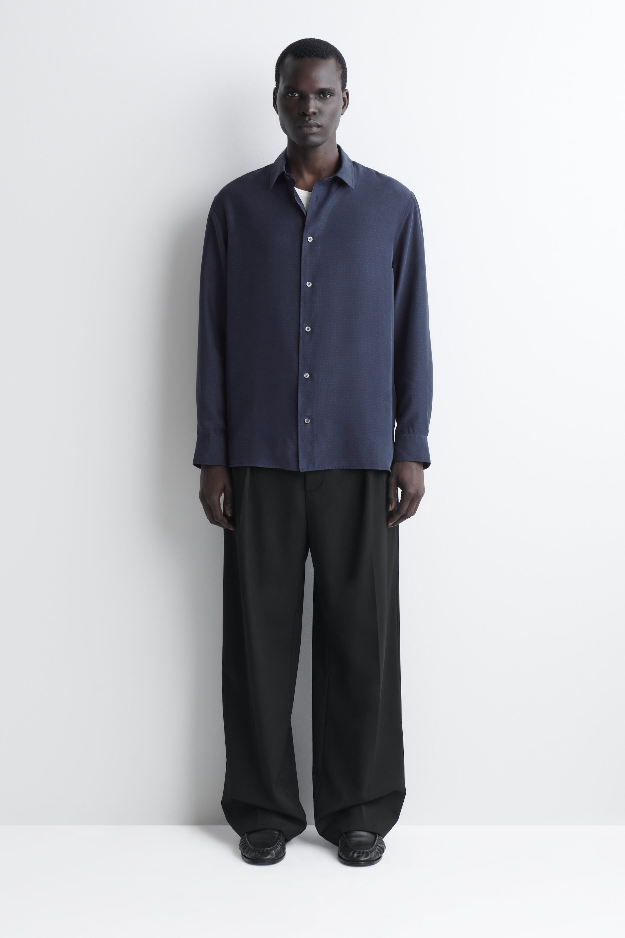 A navy blue sandwashed silk shirt is shown from the front, featuring a relaxed fit, a classic point collar, a button-front closure, and long sleeves with buttoned cuffs. The shirt is worn untucked over wide-leg black trousers, with the top button left open to reveal a hint of a white undershirt. A navy blue sandwashed silk shirt is shown from the front, featuring a relaxed fit, a classic point collar, a button-front closure, and long sleeves with buttoned cuffs. The shirt is worn untucked over wide-leg black trousers, with the top button left open to reveal a hint of a white undershirt.