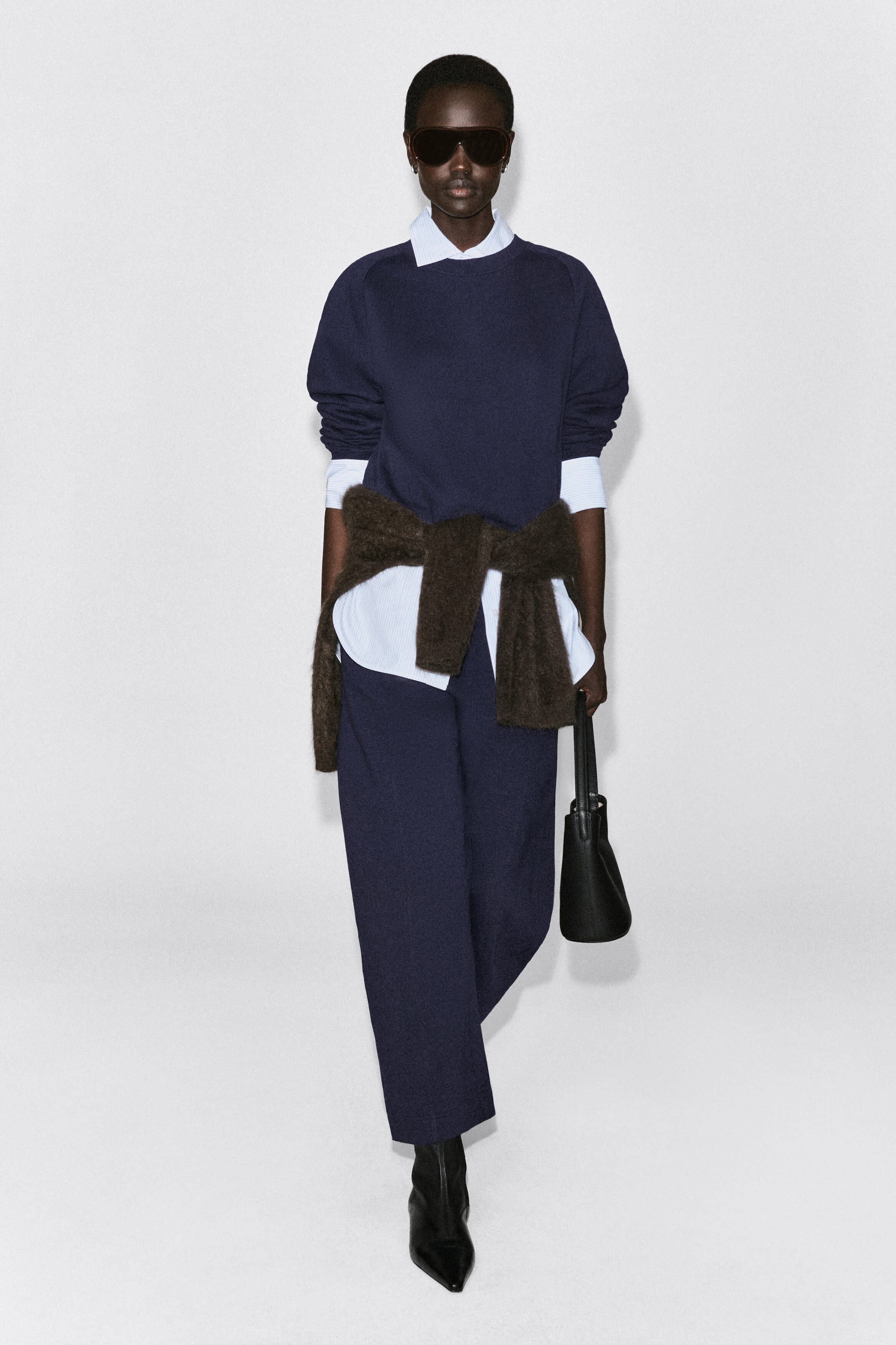 A navy blue cotton sweatshirt is shown from the front, featuring a relaxed fit, a crew neck, and long sleeves pushed up to reveal a layered light blue collared shirt. The sweatshirt is styled with matching navy trousers and a brown knit sweater tied around the waist. A navy blue cotton sweatshirt is shown from the front, featuring a relaxed fit, a crew neck, and long sleeves pushed up to reveal a layered light blue collared shirt. The sweatshirt is styled with matching navy trousers and a brown knit sweater tied around the waist.