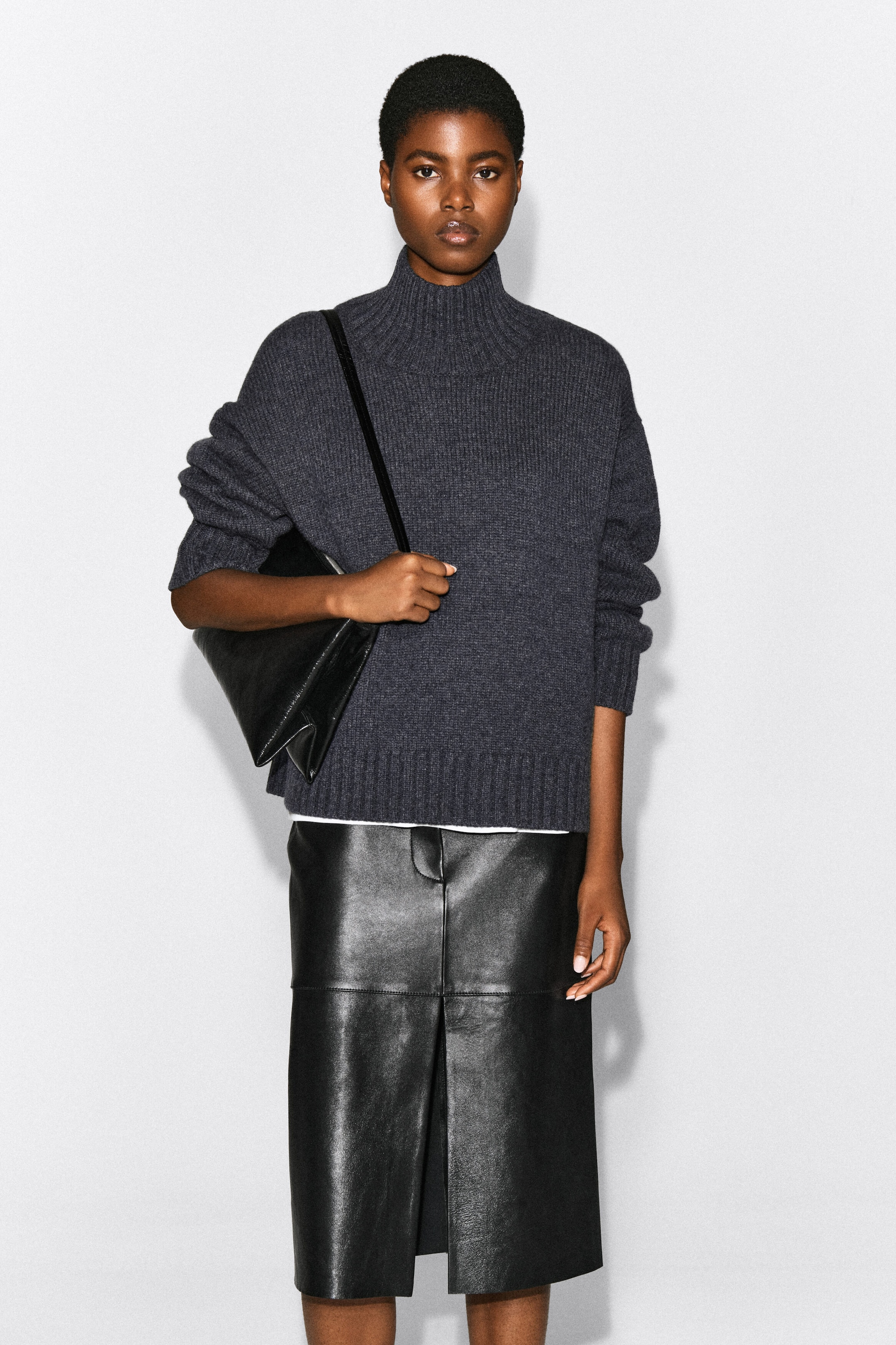 The model is wearing a chunky dark grey cashmere funnel-neck jumper, featuring a ribbed knit design and a relaxed fit. The jumper is paired with a black leather midi skirt, with the model carrying a black leather shoulder bag. The model is wearing a chunky dark grey cashmere funnel-neck jumper, featuring a ribbed knit design and a relaxed fit. The jumper is paired with a black leather midi skirt, with the model carrying a black leather shoulder bag.