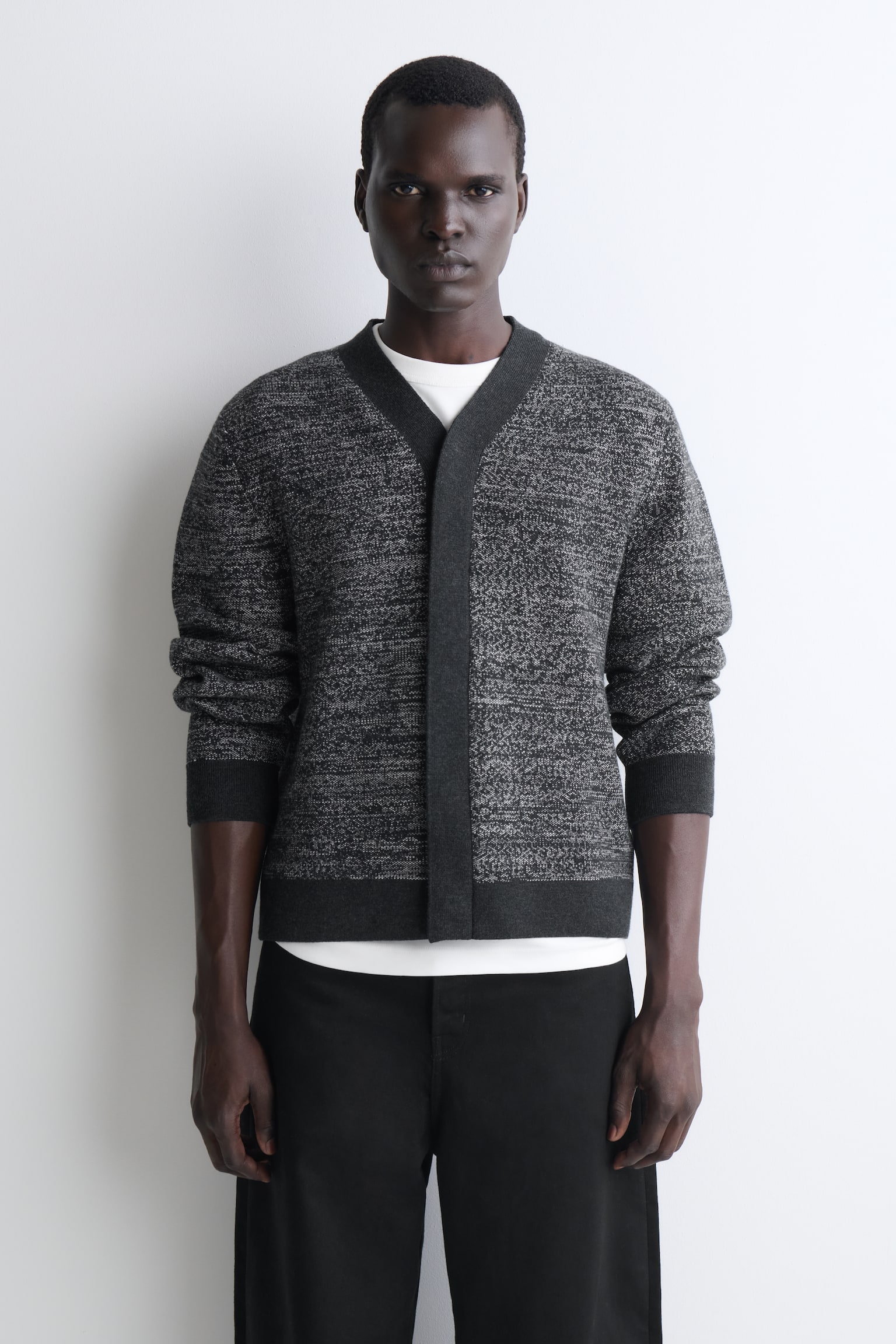 Eine melierte Strickjacke in Grau ist frontal ausgerichtet und zeigt ein strukturiertes Strickmuster in Grau und Weiss mit einer kontrastierenden, einfarbig dunkelgrauen V-Ausschnitt-Frontblende sowie Rippbündchen und Saum. Die Strickjacke wird über einem weissen T-Shirt mit Rundhalsausschnitt getragen und mit einer schwarzen Hose kombiniert, um die lockere Passform und das knopflose Design hervorzuheben. - 1