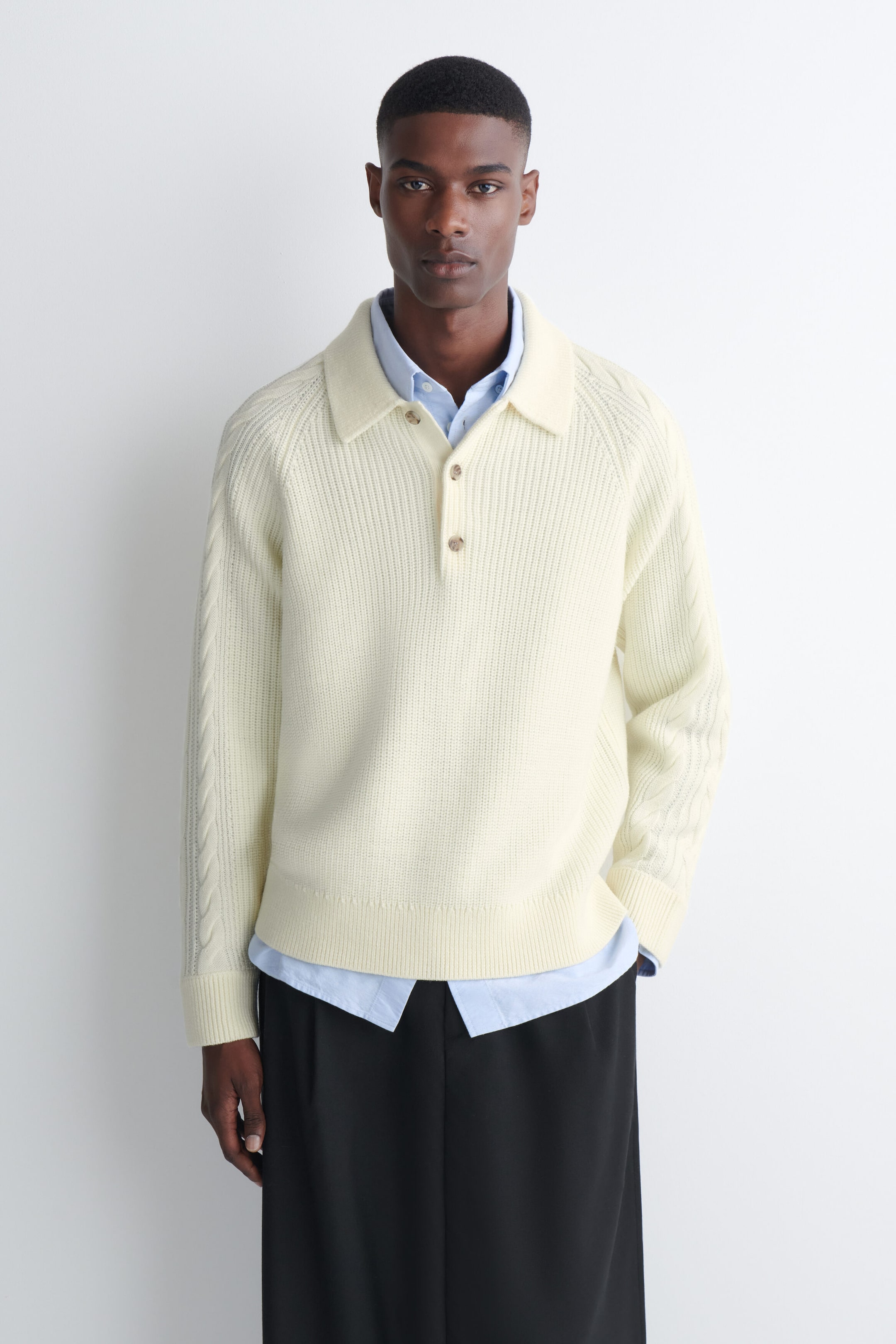 A cream-colored chunky-knit polo sweater is worn by the model, layered over a light blue collared shirt and black trousers. The sweater features a ribbed texture, a buttoned polo collar, and cable-knit detailing along the sleeves. A cream-colored chunky-knit polo sweater is worn by the model, layered over a light blue collared shirt and black trousers. The sweater features a ribbed texture, a buttoned polo collar, and cable-knit detailing along the sleeves.