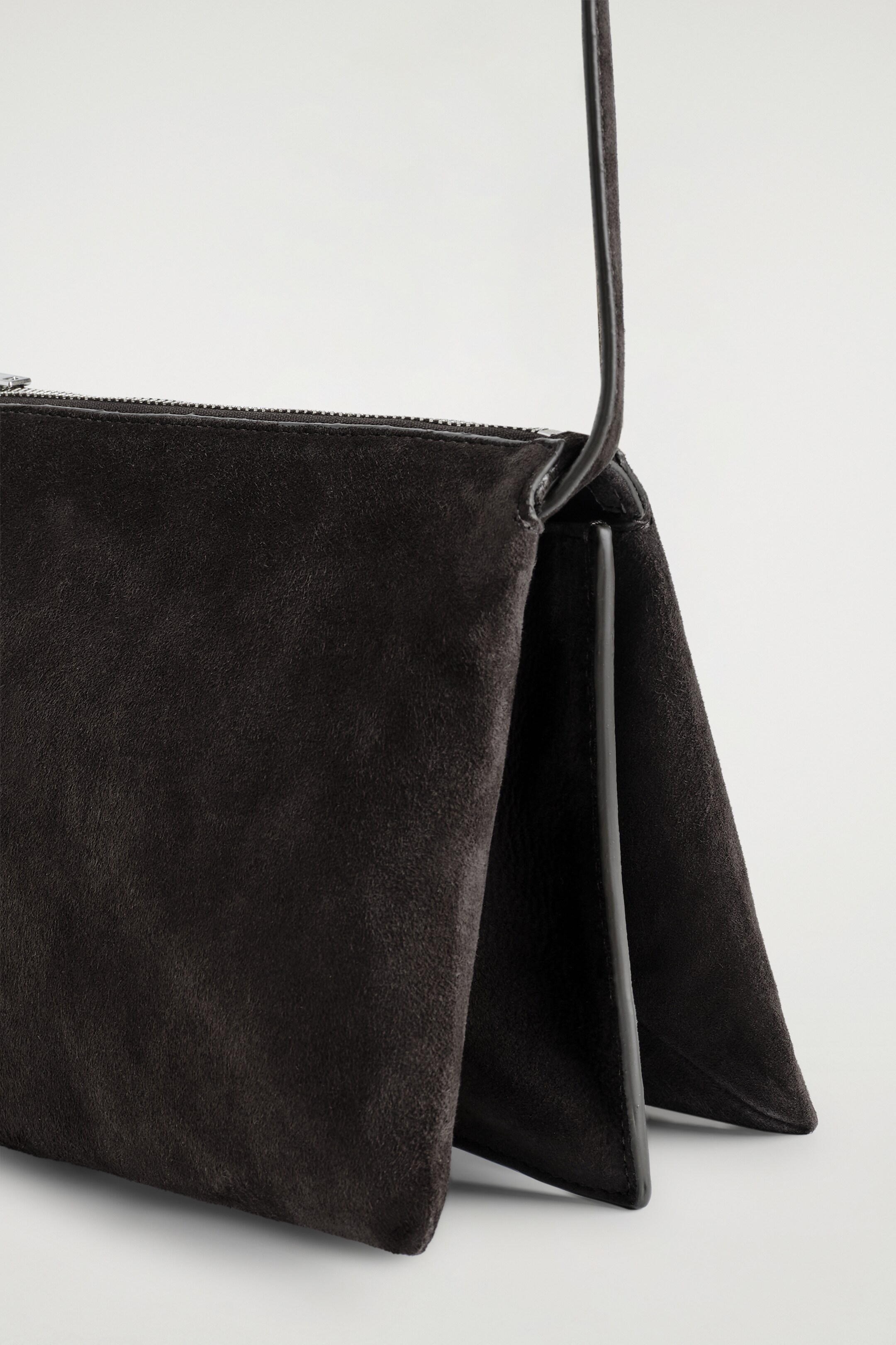 Visualizza immagine più grande: La TROVE CROSSBODY BAG - SUEDE mostra una borsa in pelle scamosciata scura, leggermente inclinata a sinistra, che rivela una profonda costruzione piegata sul lato destro. Presenta una chiusura con cerniera superiore e una sottile tracolla che si estende verso l'alto dalla parte superiore destra.