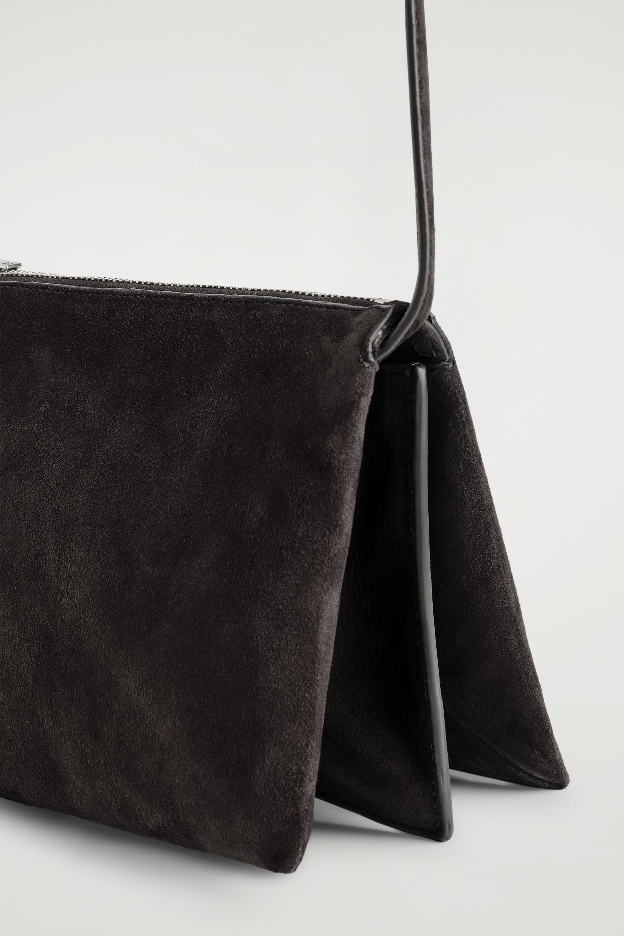 Agrandir l'image: TROVE CROSSBODY BAG - SUEDE montre un sac en daim foncé, légèrement incliné vers la gauche, révélant une construction profonde et pliée sur son côté droit. Il est doté d'une fermeture éclair sur le dessus et d'une fine bandoulière qui s'étend vers le haut depuis le coin supérieur droit.