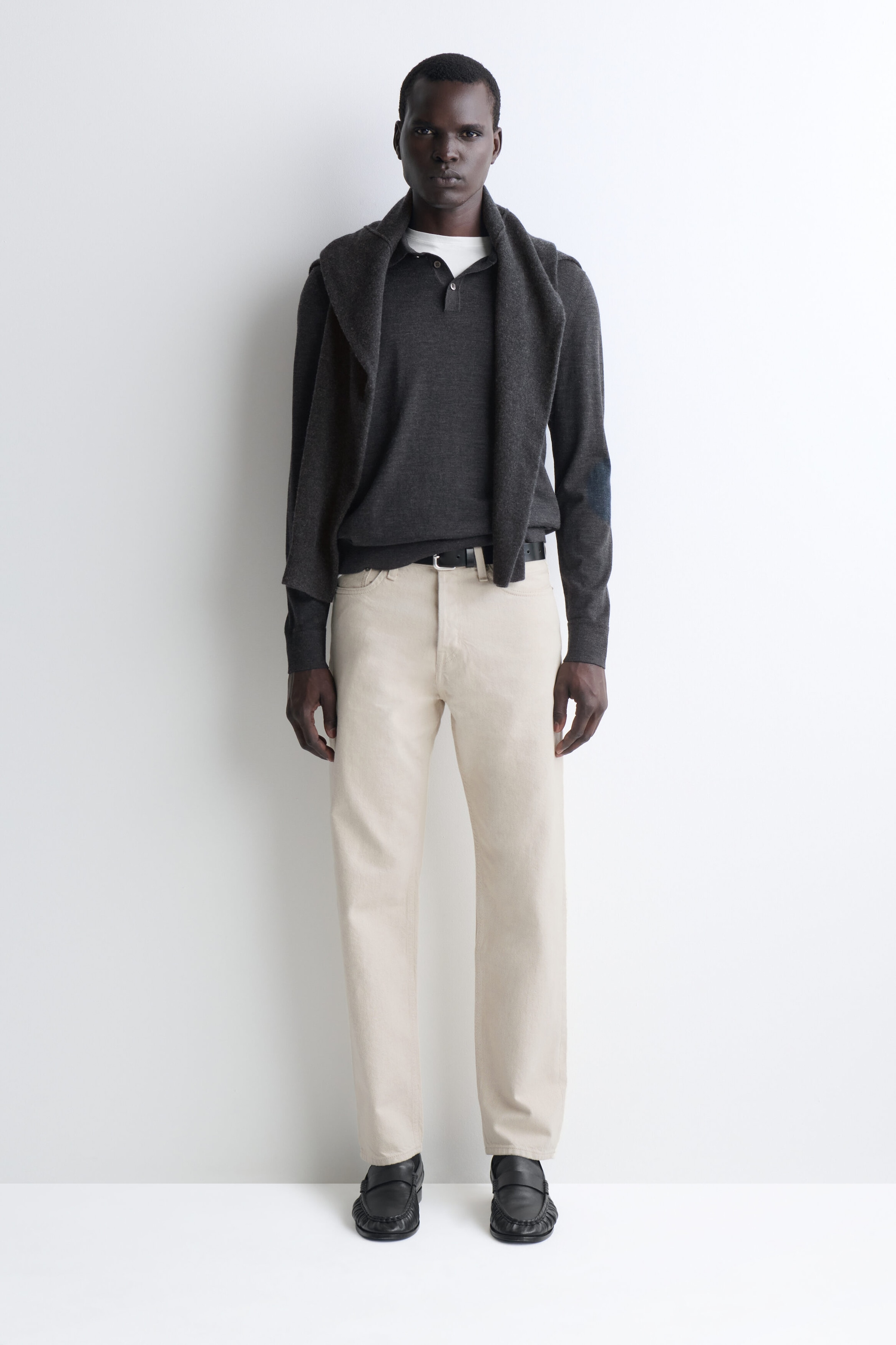 Ein Model steht frontal und trägt ein dunkelgraues langärmliges Strick-Poloshirt mit Knopfleiste, kombiniert mit cremefarbenen, gerade geschnittenen Hosen. Ein passender dunkelgrauer Strickpullover liegt über seinen Schultern, und die Hose ist mit einem schwarzen Gürtel fixiert. Ein Model steht frontal und trägt ein dunkelgraues langärmliges Strick-Poloshirt mit Knopfleiste, kombiniert mit cremefarbenen, gerade geschnittenen Hosen. Ein passender dunkelgrauer Strickpullover liegt über seinen Schultern, und die Hose ist mit einem schwarzen Gürtel fixiert.